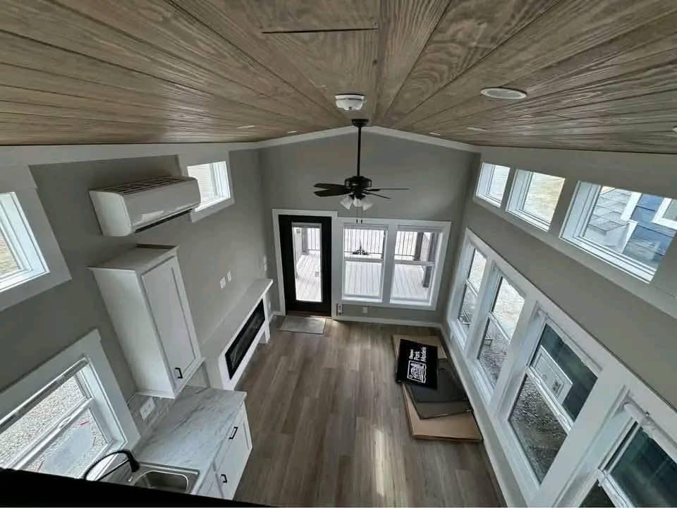 Spacious, light-filled interior of a modern tiny house with a high, wood-paneled vaulted ceiling, a black ceiling fan, and recessed lighting, featuring a gray-painted wall with a white built-in cabinet, a sleek fireplace, and a wall-mounted air conditioning unit, complemented by a kitchen area with white marble countertops, a stainless steel sink, and a black faucet, all set against a backdrop of numerous large windows offering ample natural light and views of the exterior.