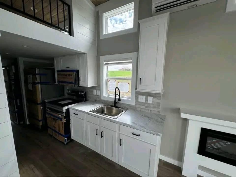 Modern tiny home kitchen with white cabinets, marble countertops, stainless steel sink, stove, refrigerator, microwave, fireplace, and loft access, showcasing compact living design.