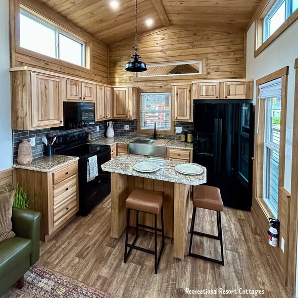 Cozy rustic kitchen with wooden cabinetry and vaulted ceiling. Granite island with two stools, black appliances, and warm ambient lighting.