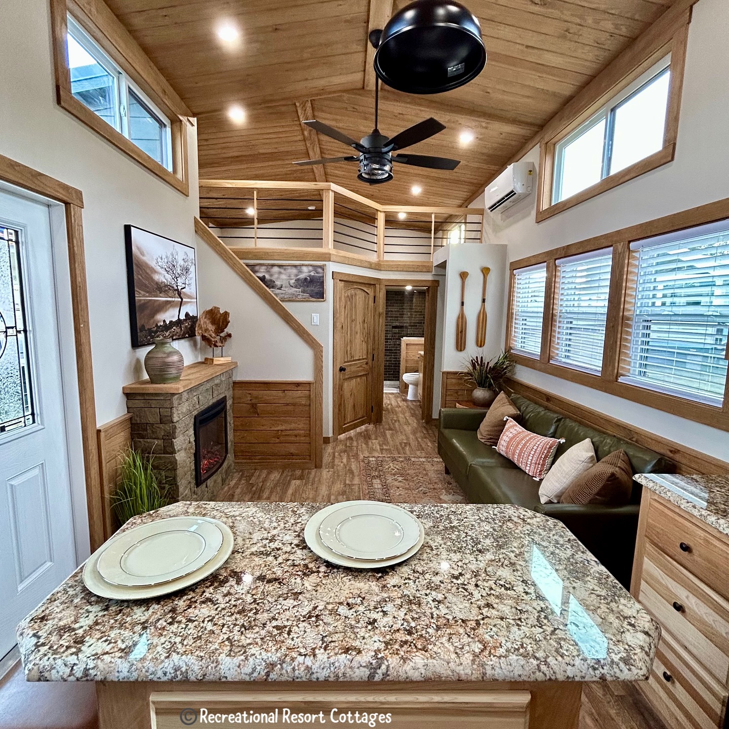 Warm, cozy interior of a tiny house with wood accents. Features a loft, stone fireplace, green sofa, granite kitchen island, and ceiling fan.