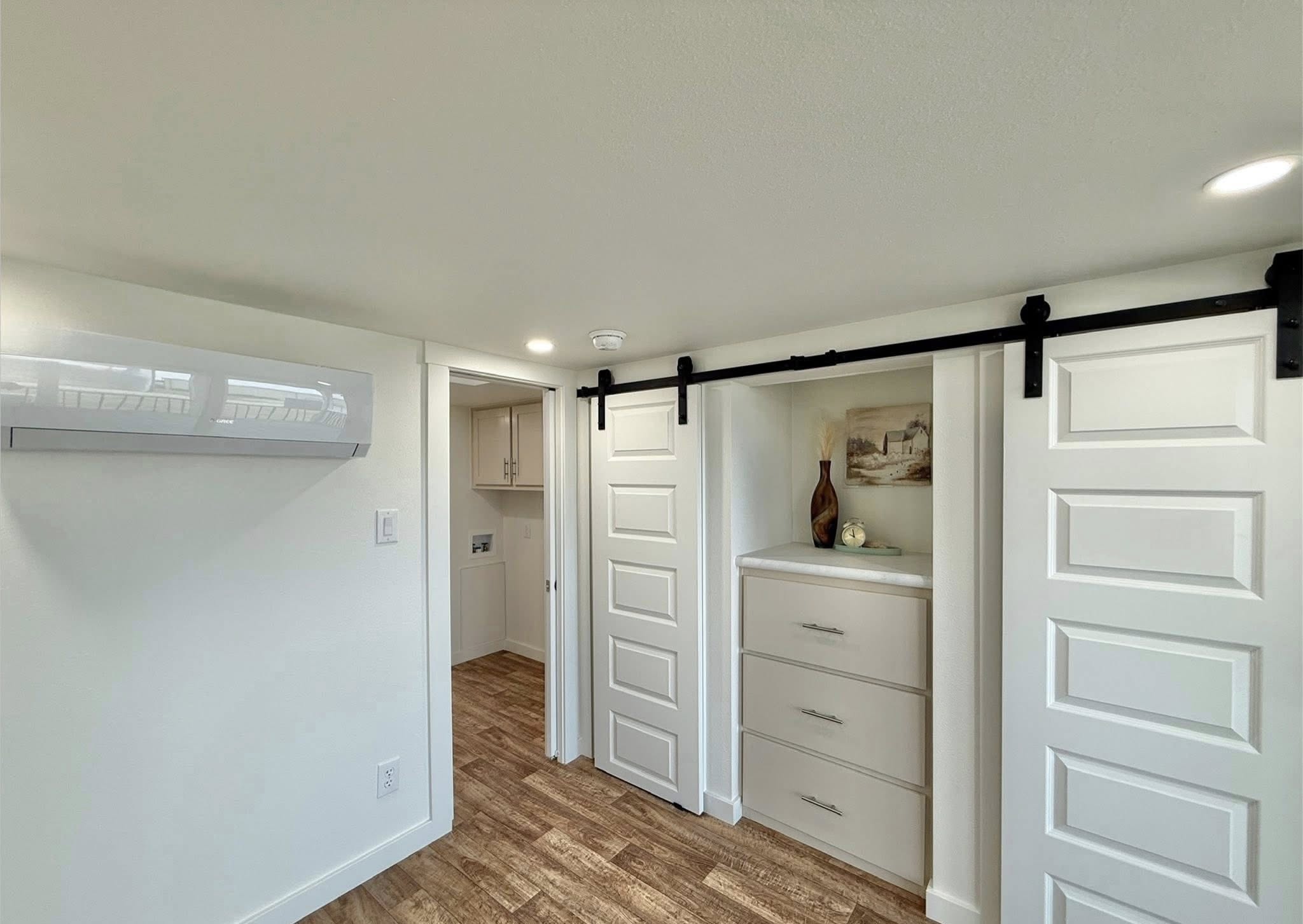 Modern tiny house interior with a white mini-split AC unit, a hallway leading to a laundry area with cabinets and a washer-dryer, a white paneled door on a black barn door track, and a built-in dresser with drawers next to a decorative vase and artwork, all on a wood-look floor.