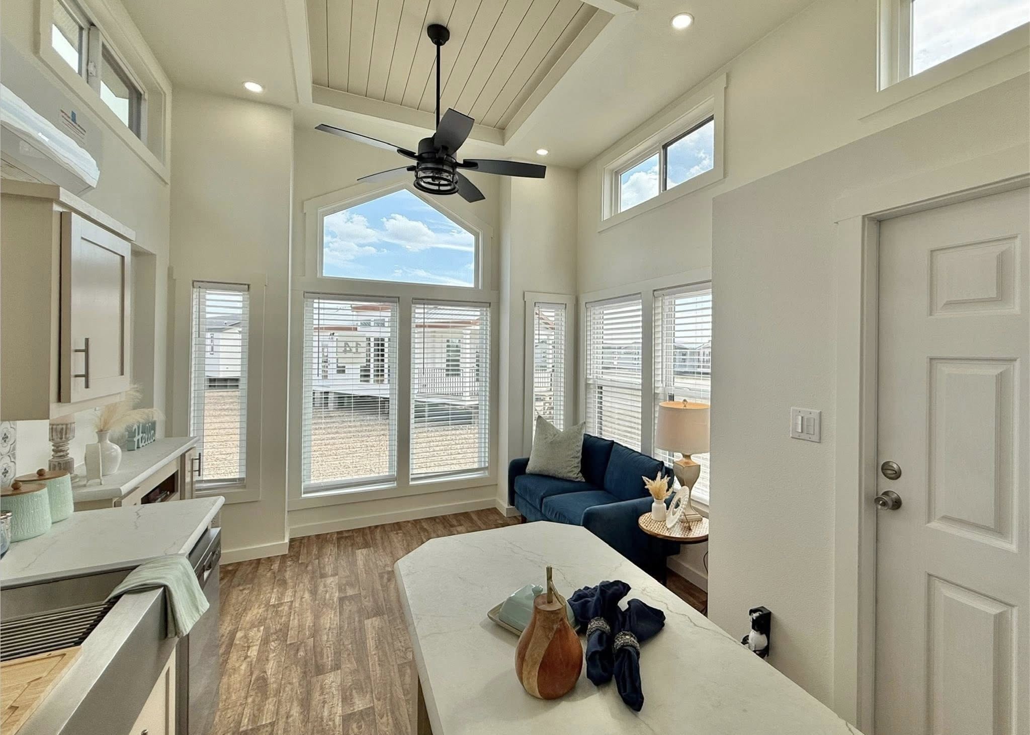 Bright and airy modern tiny house interior featuring a spacious open-concept living area with a vaulted ceiling, a black ceiling fan, a large geometric window, a comfortable blue sofa, and a sleek white kitchen with light wood-look flooring.