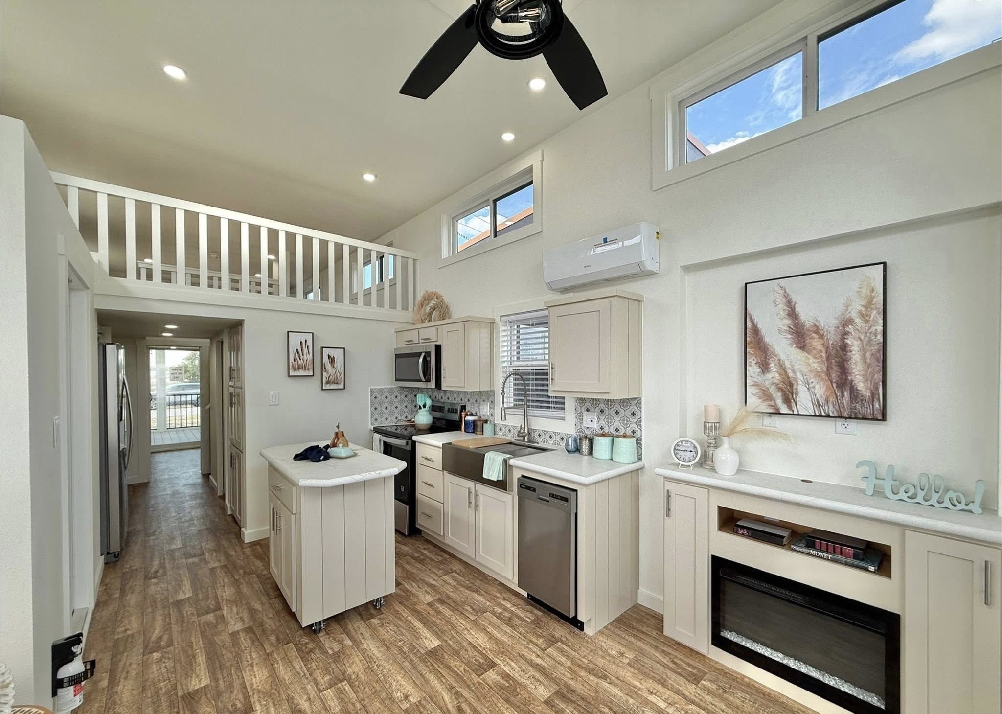 Spacious interior of a modern tiny house showcasing a bright and open living area with a loft bedroom, a full kitchen with light-colored cabinets and stainless steel appliances, a cozy fireplace, and a long hallway leading to the entrance, all complemented by warm wood-look flooring and ample natural light from large windows.