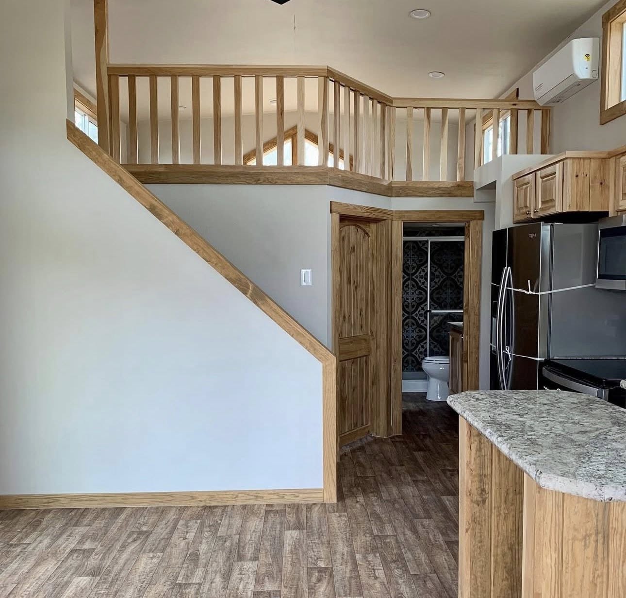 Spacious open-concept modern farmhouse tiny home interior featuring dramatic lofted upper level with wooden balustrade railing and exposed beams, curved wooden staircase with light pine handrails leading to cozy loft bedroom overlook, vaulted ceiling with recessed lighting and black ceiling fan, neutral light gray walls, warm medium-tone luxury vinyl plank flooring, light pine shaker kitchen cabinets with granite countertops, stainless steel appliances including side-by-side refrigerator and electric range, patterned Moroccan tile backsplash, high clerestory windows flooding abundant natural light, open layout flowing to bathroom entrance with decorative dark tile shower visible, airy bright multi-level design perfect for compact tiny house living, modular cottage retreats, or family-friendly vacation homes.