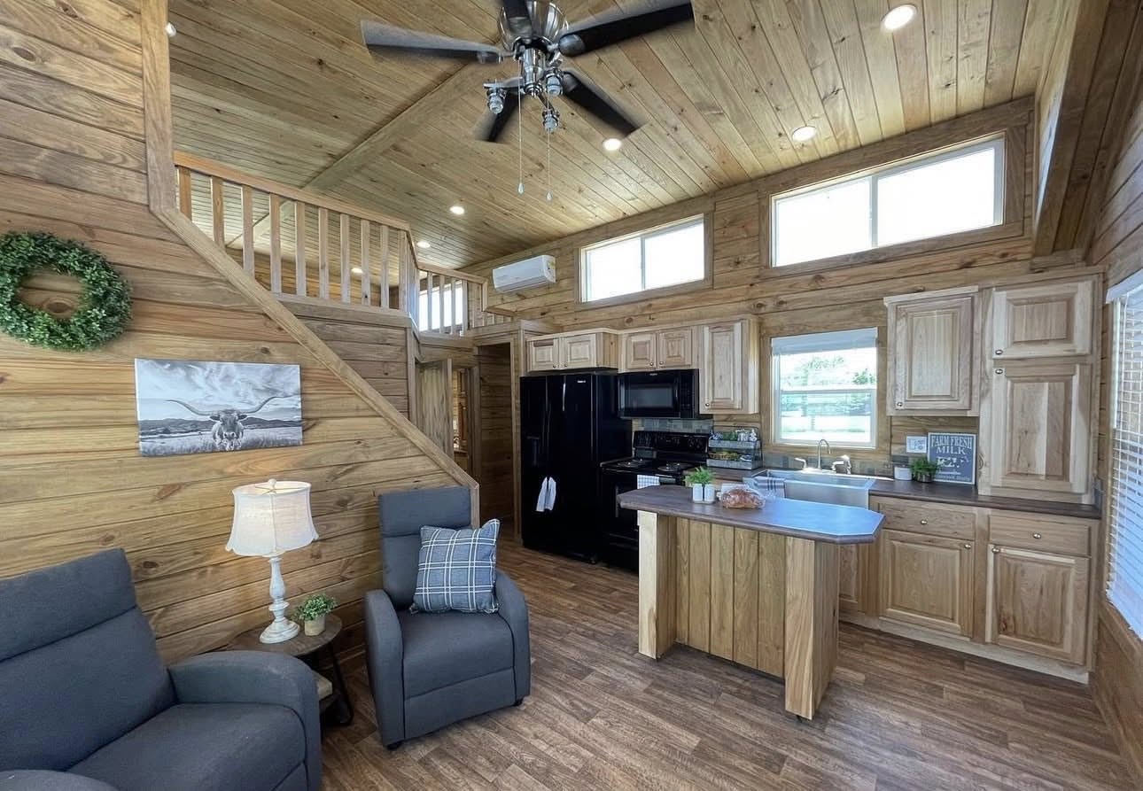 Cozy cabin interior with wooden walls and ceiling. Features include a modern kitchen with black appliances, a loft, gray recliners, and a rustic atmosphere.