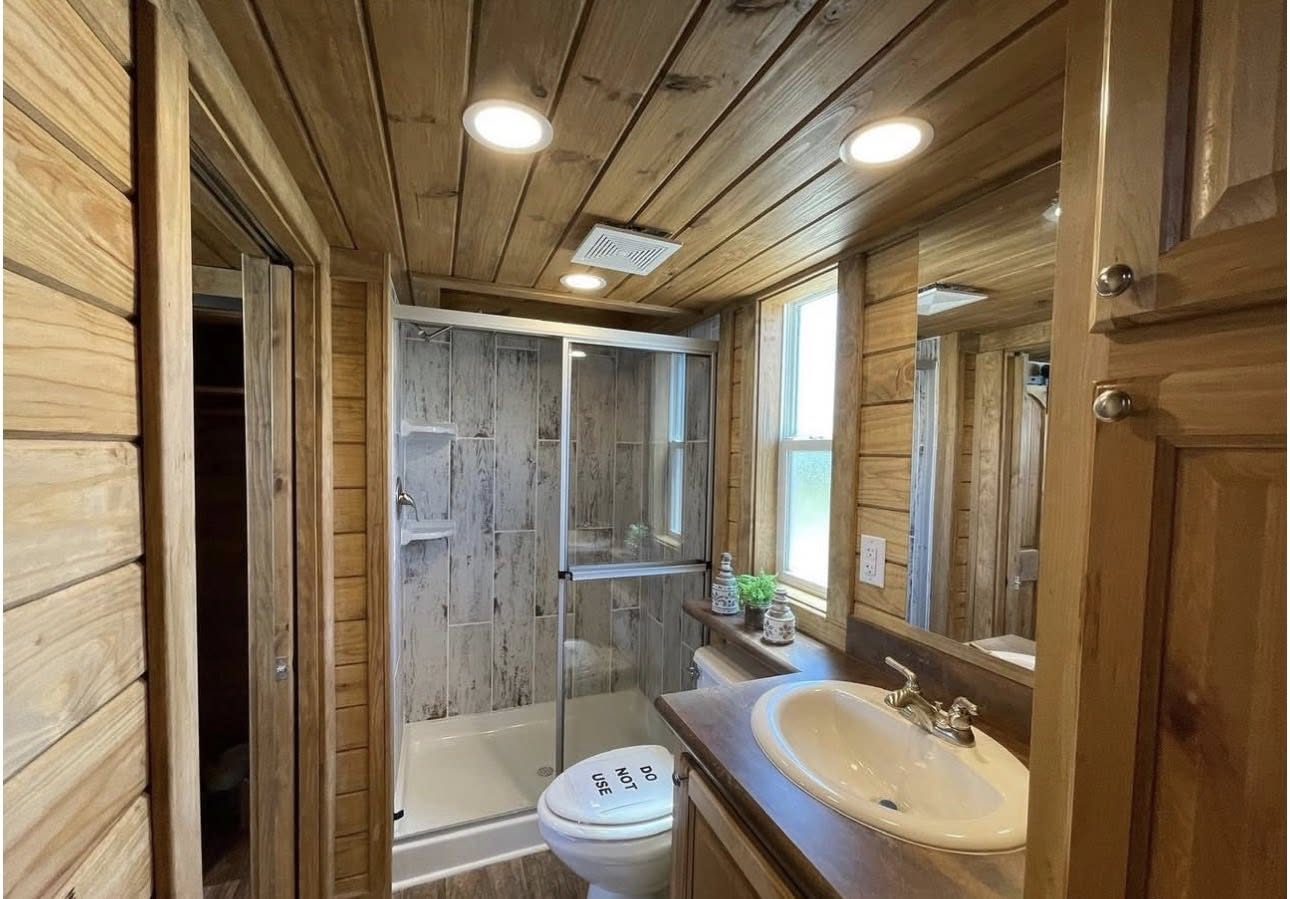 Rustic bathroom with wood paneling, a glass shower, and a vanity with a white sink and mirror. Natural light streams in from a window, creating a cozy feel.