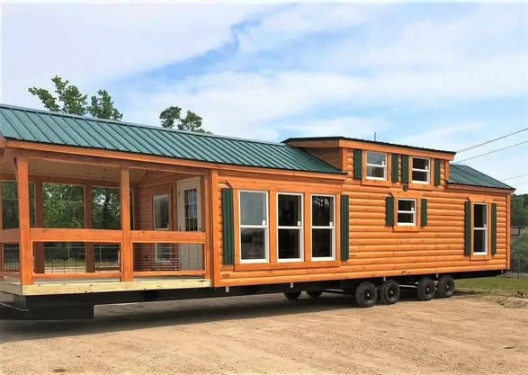 A mobile cabin with a wooden exterior and a green metal roof sits on a dirt road. It features large windows and a covered porch, conveying a cozy, rustic feel.