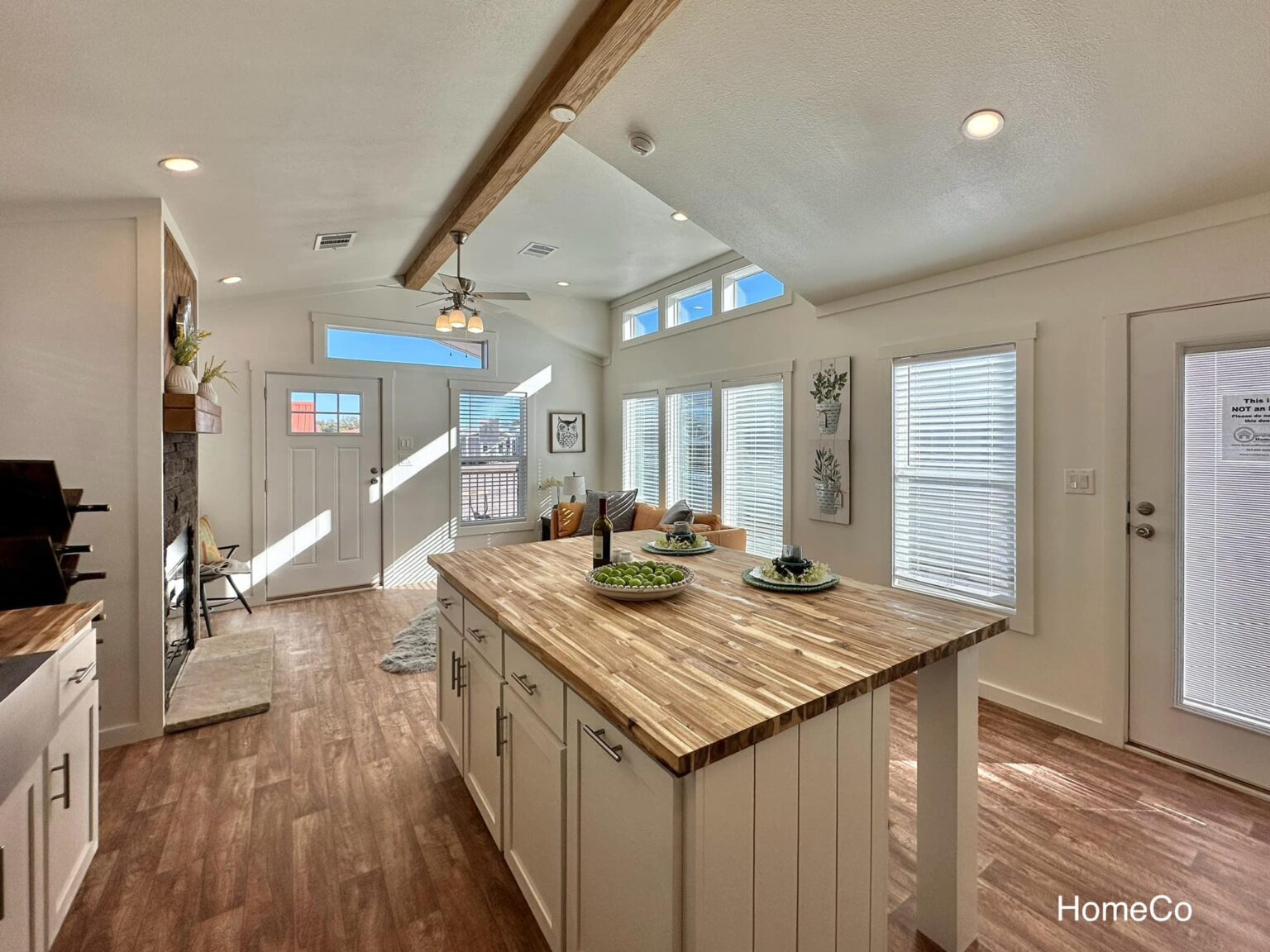 Modern luxury farmhouse-style park model tiny home interior featuring an expansive open-concept living area with a large butcher block kitchen island, high vaulted ceilings with a rustic natural wood beam, and a contemporary five-blade ceiling fan. The sophisticated architectural design showcases bright white shiplap walls, premium wide-plank wood flooring, and a stone-accented fireplace that adds a cozy touch to the minimalist aesthetic. Large energy-efficient windows with white blinds flood the space with natural light, highlighting the gourmet kitchen's white cabinetry and high-end stainless steel appliances. This professional "full rank" SEO-optimized small-space living solution is an ideal showcase for custom-built prefab homes, luxury vacation cottages, or modern backyard accessory dwelling units (ADU) designed for maximum style, comfort, and functional elegance.