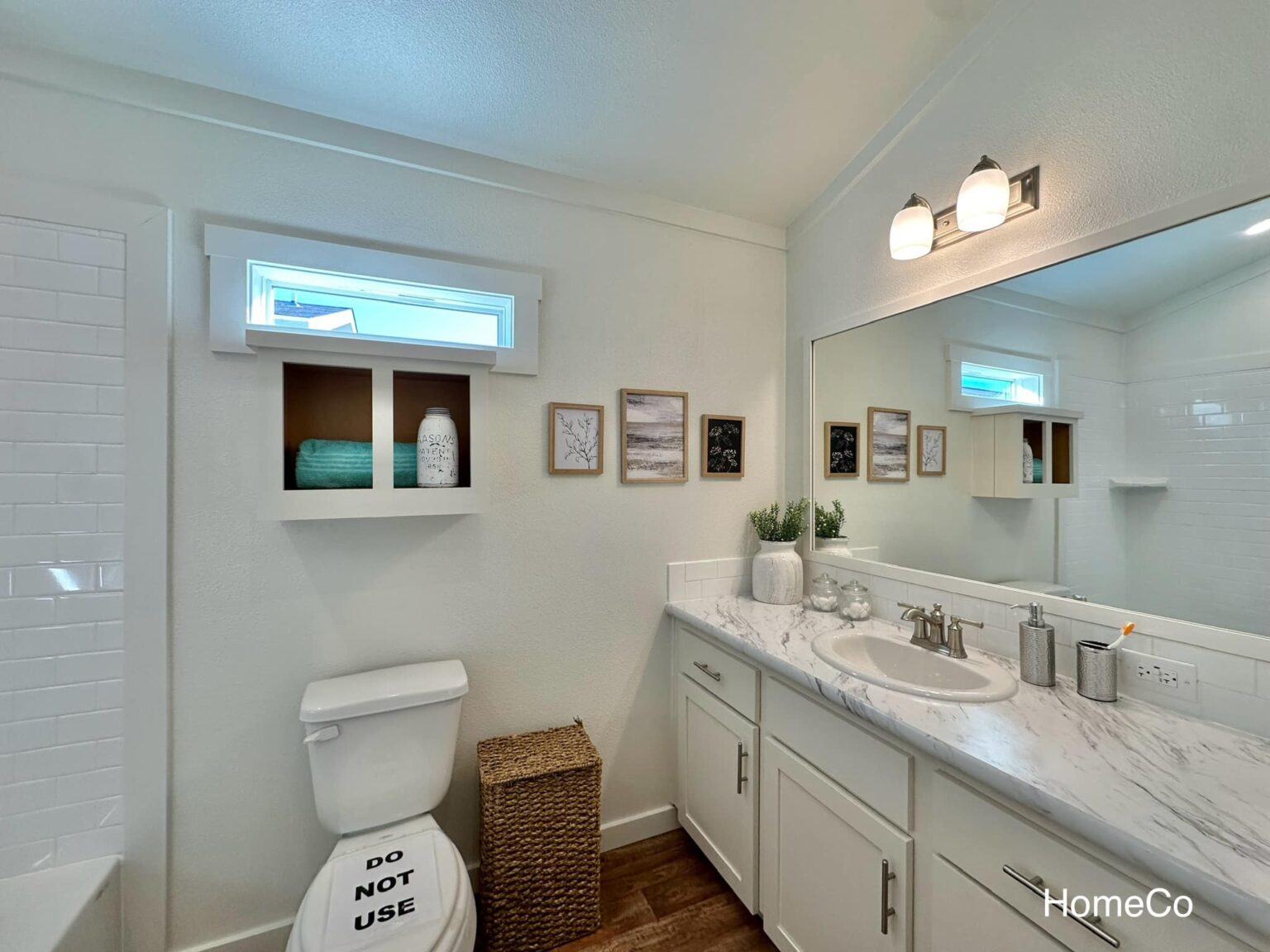 Modern luxury farmhouse-style bathroom in a park model tiny home featuring a crisp white vanity with a marble-patterned countertop, a large rectangular mirror with contemporary vanity lighting, and elegant silver fixtures. The sophisticated architectural design showcases clean white walls, a built-in recessed storage nook with a decorative mason jar, and premium wide-plank wood flooring that complements the minimalist aesthetic. A bright energy-efficient transom window floods the space with natural light, highlighting the professional "full rank" SEO-optimized interior which includes a walk-in tiled shower and stylish decor, making it an ideal inspiration for custom-built prefab homes, luxury vacation cottages, or modern backyard accessory dwelling units (ADU) designed for maximum comfort and high-end appeal.