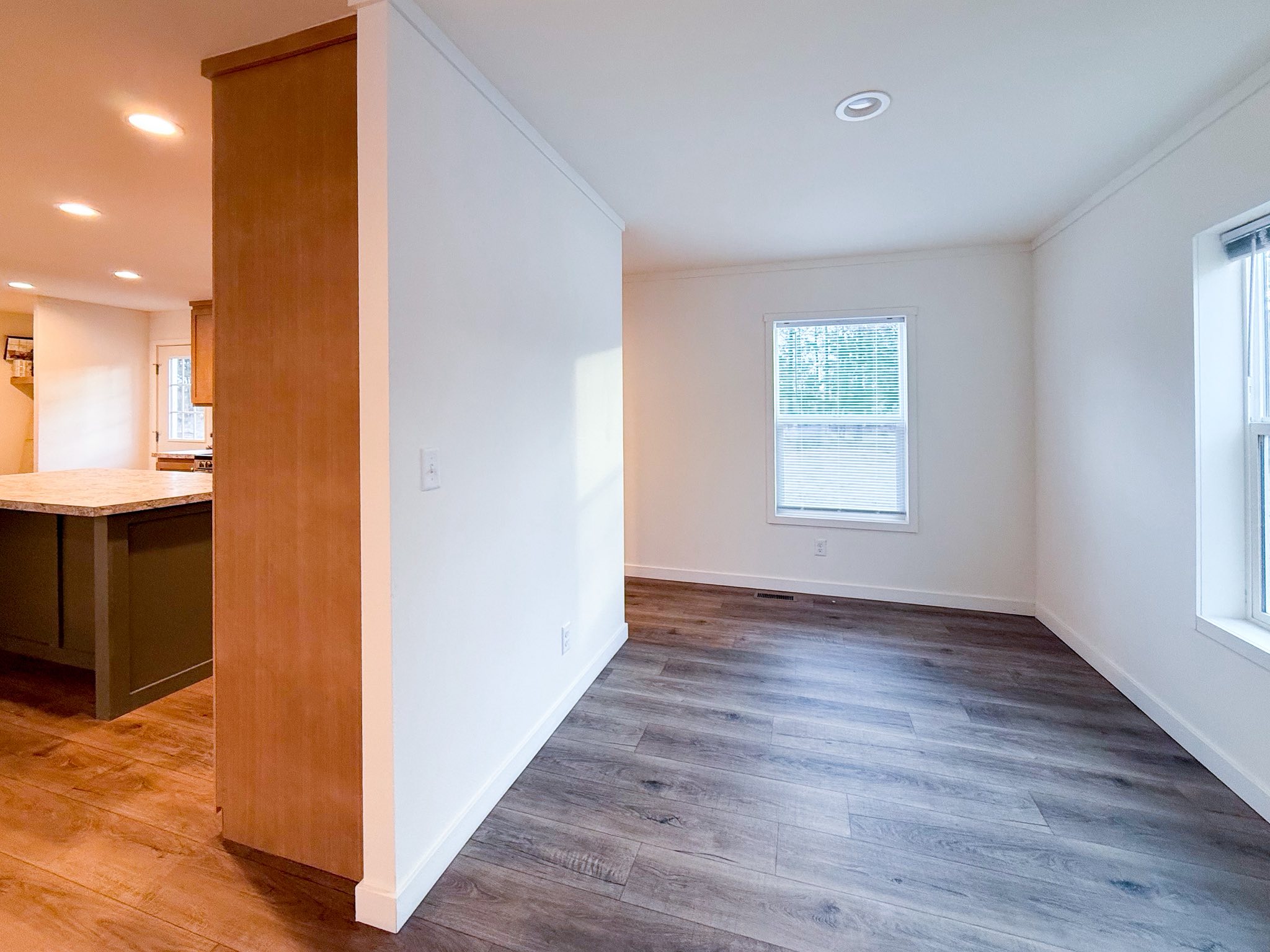 Modern luxury modular home interior showcasing a bright, empty open-concept room with premium wide-plank grey wood flooring and crisp white walls. The sophisticated architectural layout features an adjacent kitchen with a large dark olive green island and marble-style countertops, illuminated by recessed LED lighting to highlight a clean, minimalist aesthetic perfect for a high-end prefab house or contemporary custom-built small home.