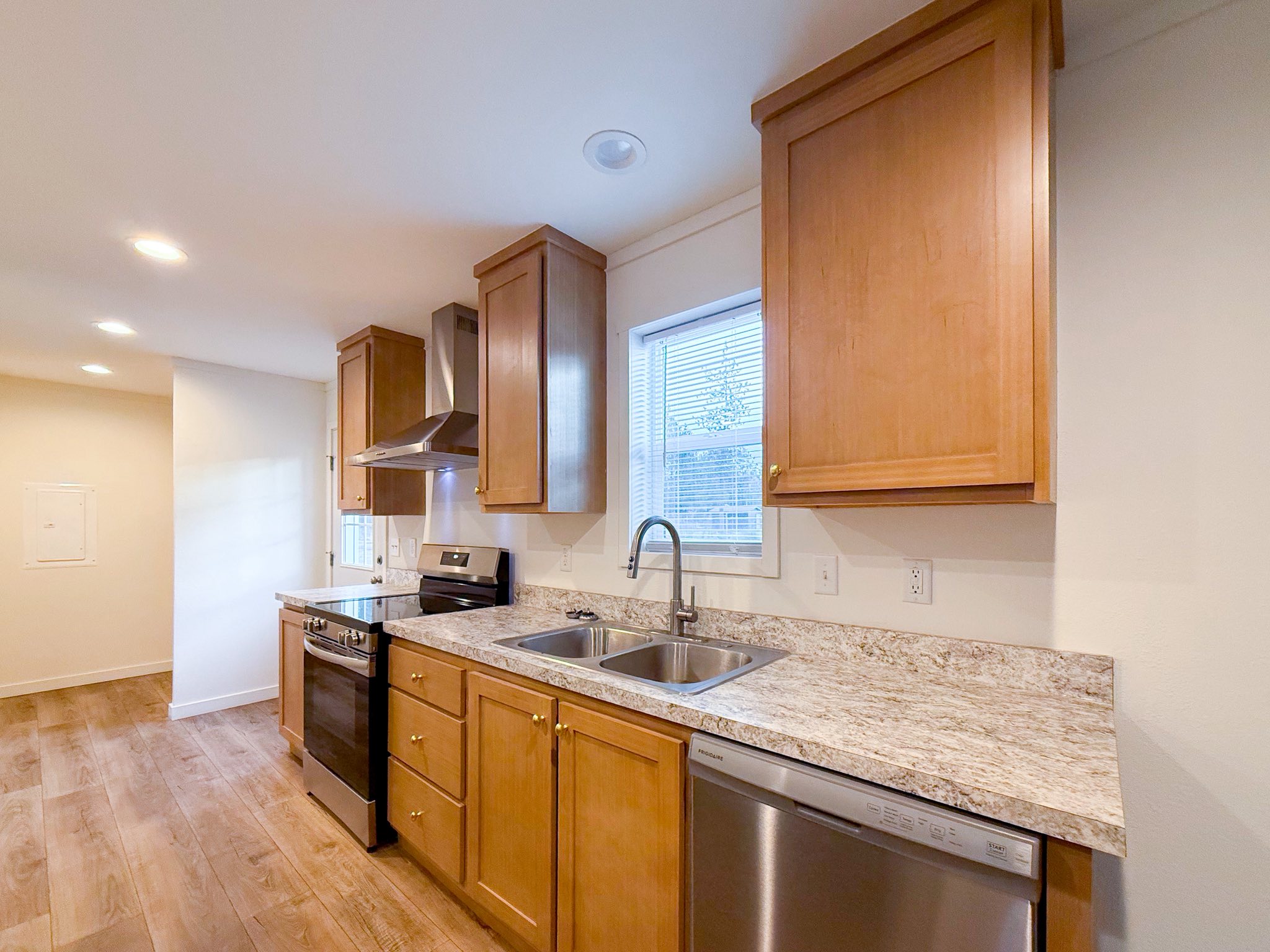 Modern luxury modular home kitchen interior featuring warm wood cabinetry, a granite-style laminate countertop with a double stainless steel sink, and high-end stainless steel Frigidaire appliances. The bright, open-concept space includes energy-efficient LED recessed lighting, a modern range hood, and premium wide-plank flooring, creating a sophisticated and functional culinary area for a contemporary prefab house or custom-built small home.