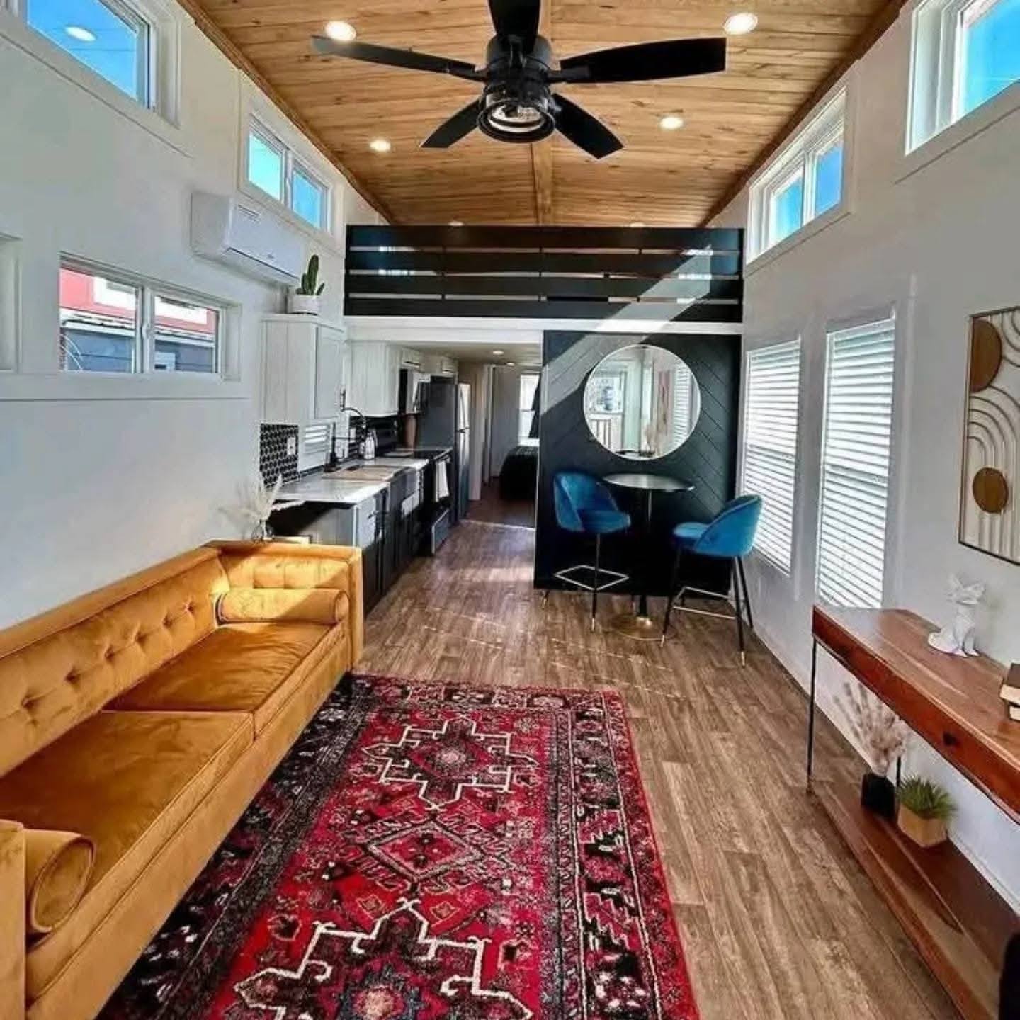 Stylish tiny home interior with wooden ceiling and flooring, mustard sofa, vibrant red patterned rug, modern dining nook, and black kitchen. Bright and cozy.