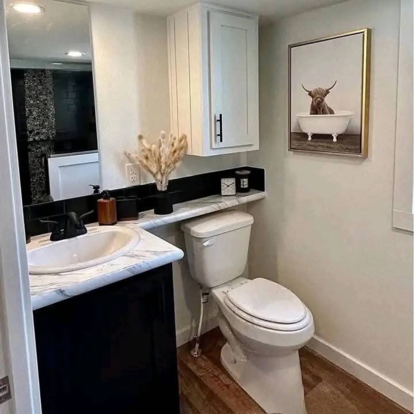 Small, modern bathroom with a white toilet, sink, and cabinet. A humorous cow-in-bathtub artwork hangs on white walls. Warm, inviting tones.