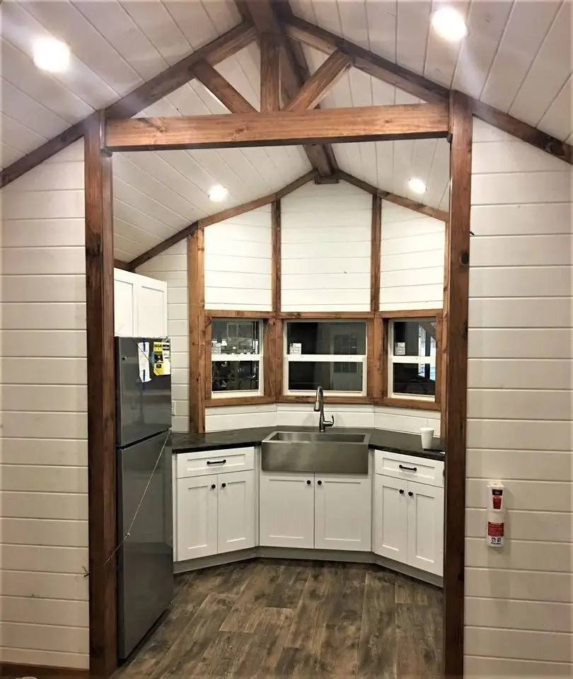 Cozy kitchen with a wooden vaulted ceiling and beams. It features a stainless steel sink, white cabinets, and a fridge. Warm, inviting atmosphere.