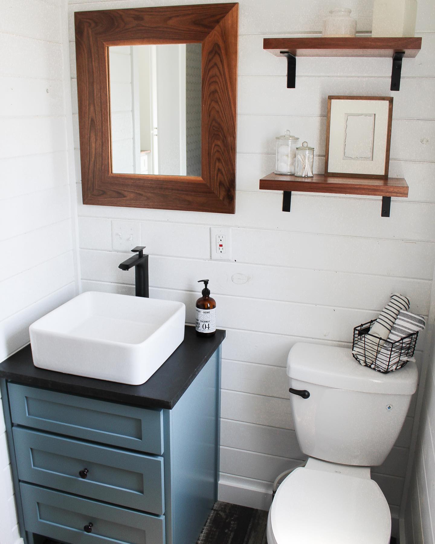 A modern, minimalist bathroom featuring a white square vessel sink with a matte black faucet mounted on a blue vanity with black countertop, a white toilet with a black metal basket holding rolled striped towels, rustic wooden floating shelves with glass jars and a framed art piece, and a large wooden-framed mirror against white shiplap walls, creating a clean, cozy, and stylish farmhouse aesthetic.