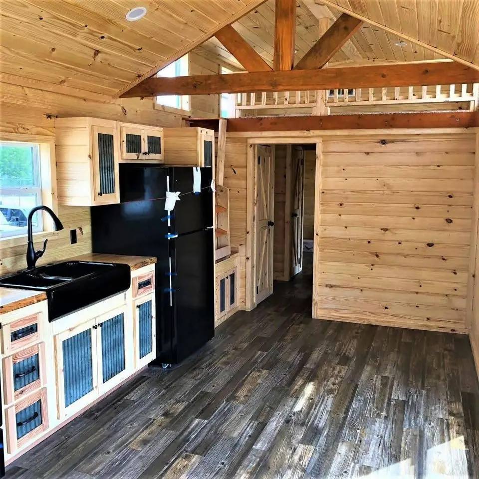 Spacious tiny house kitchen with natural wood paneling, black sink, and refrigerator. Open layout with rustic beams and loft, evoking a cozy vibe.