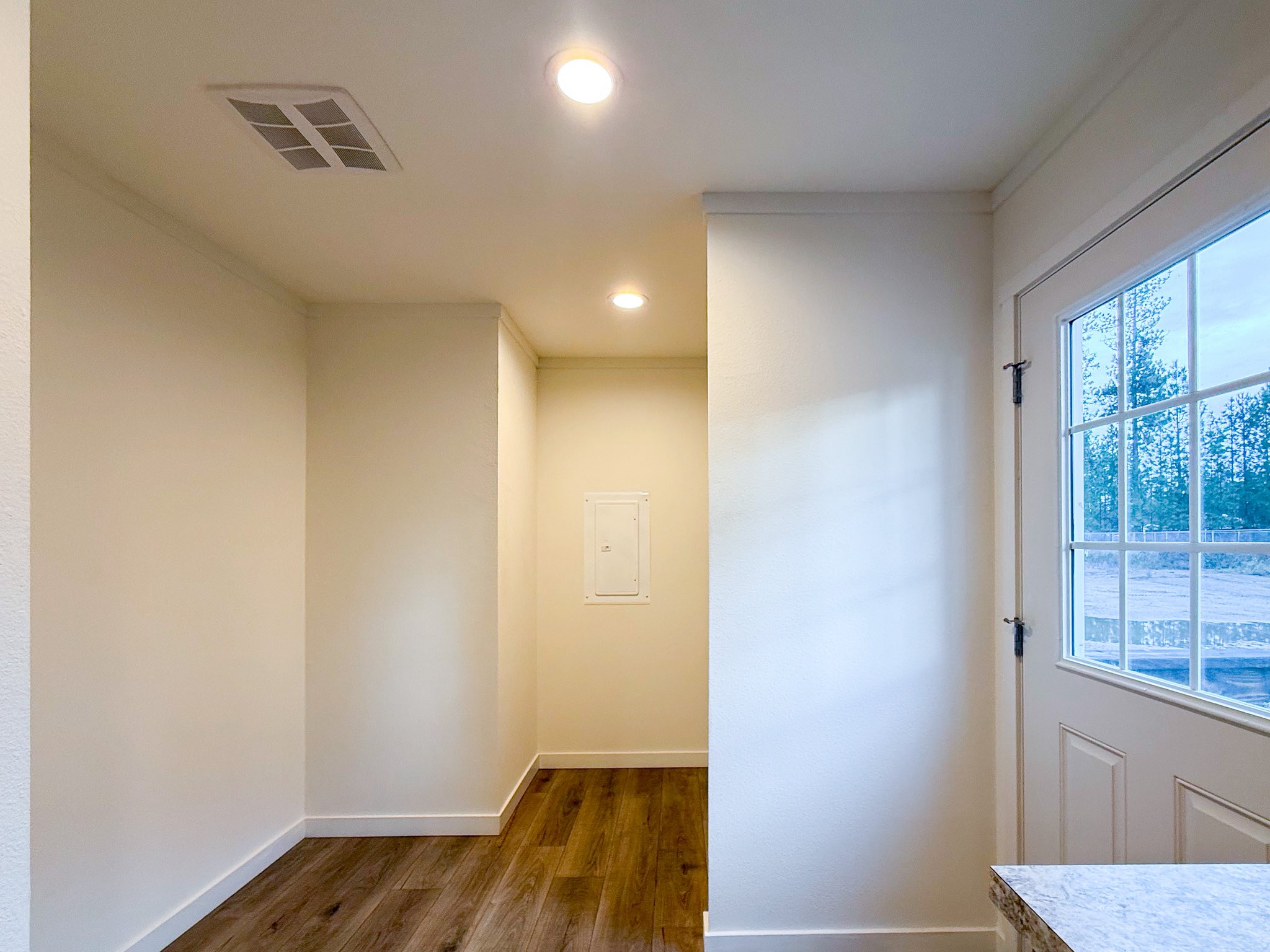 Modern luxury modular home interior featuring a bright, minimalist entryway and hallway with crisp white walls, premium wide-plank wood flooring, and energy-efficient LED recessed lighting. The clean architectural design showcases a glimpse of a contemporary kitchen with stone-patterned countertops and warm wood cabinetry, highlighting a spacious and functional floor plan perfect for high-end prefab living or a custom-built small home aesthetic.