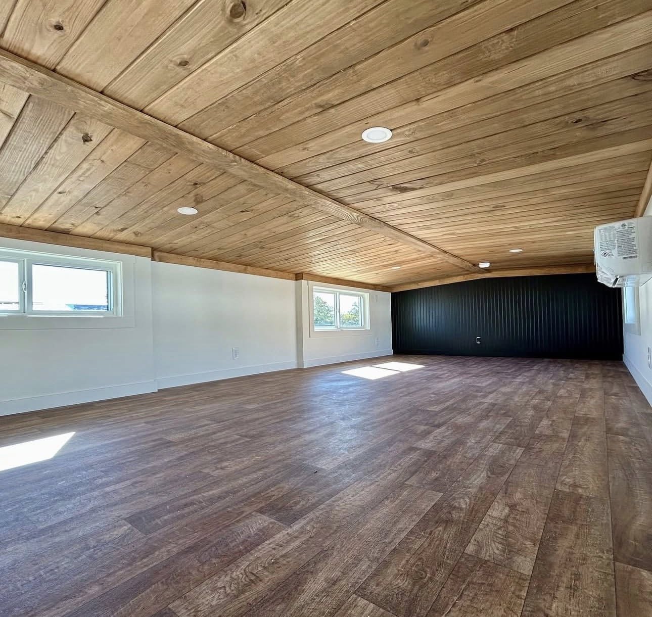 Spacious attic room with wood-paneled ceiling, laminate flooring, and small windows. Light and airy atmosphere; white and black walls enhance modern feel.
