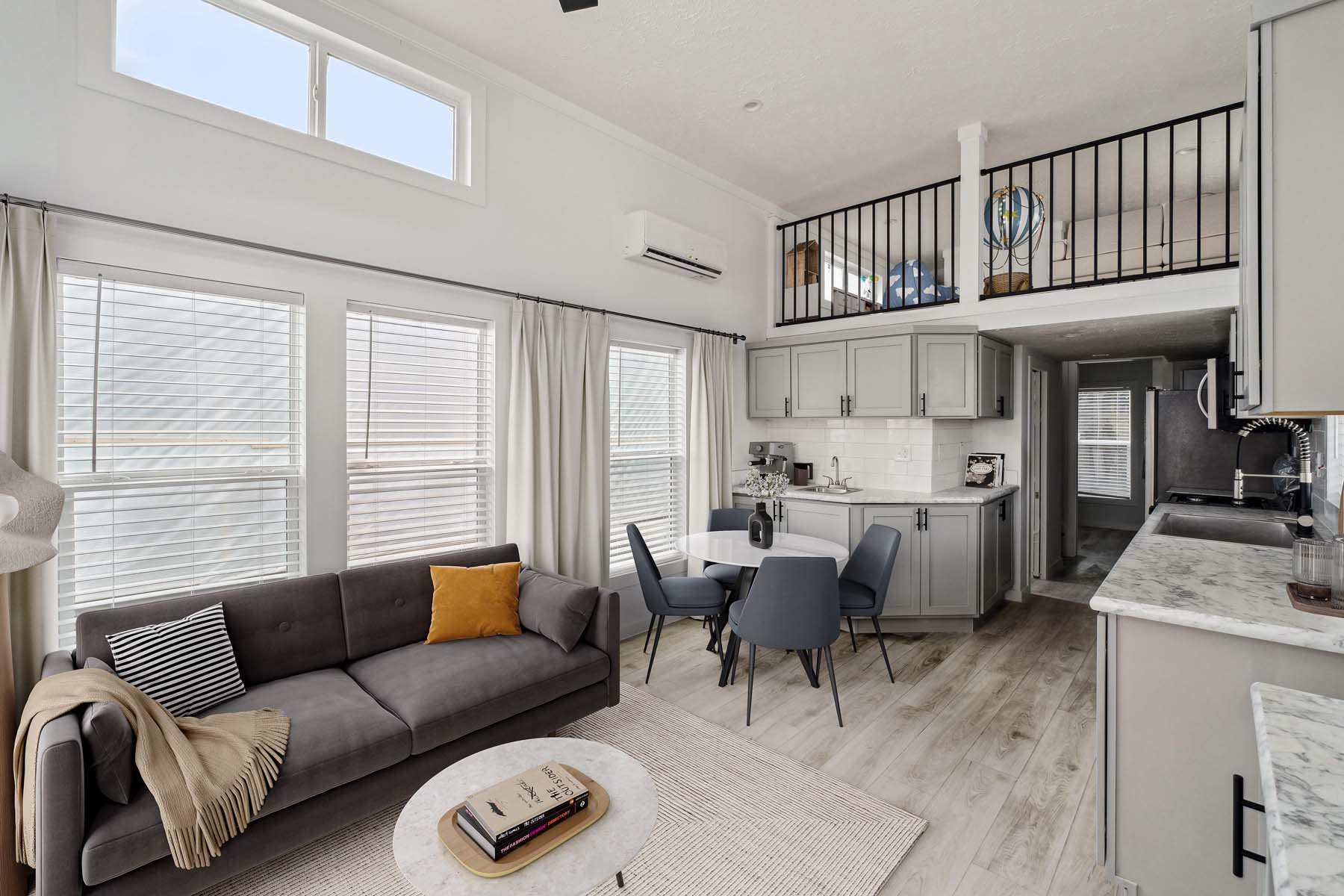 Modern living room with a gray sofa, yellow and striped pillows, and a coffee table. Open kitchen with gray cabinets, dining area, and a loft above. Bright and cozy ambiance.