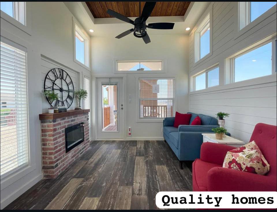Cozy modern living room with high ceilings, wood floors, and large windows. Features a brick fireplace, blue sofa, red armchair, and a wall clock.