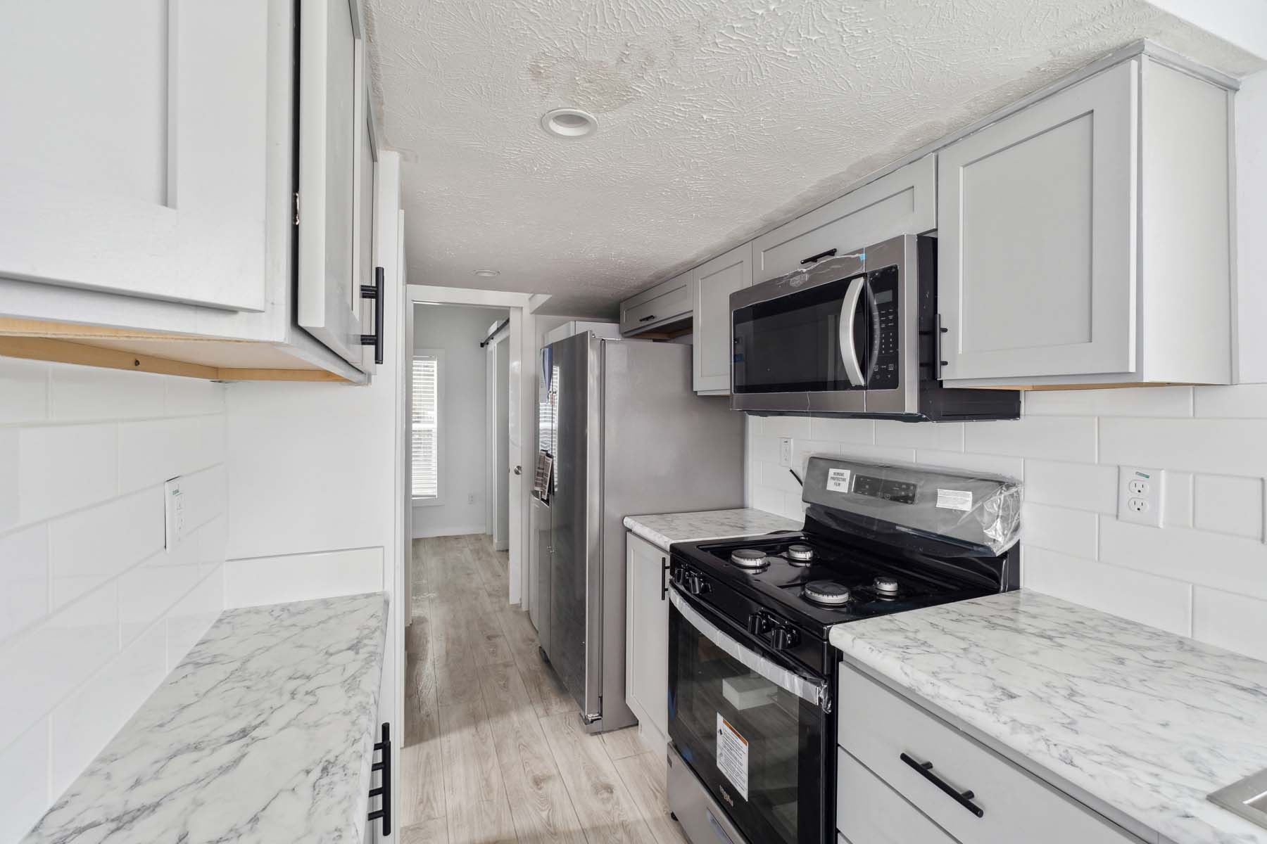 Compact modern kitchen with white cabinets, marble countertops, and a gray laminate floor. Stainless steel refrigerator, oven, and microwave. Bright natural light.