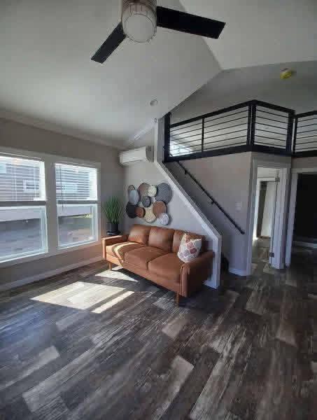 Modern living room with brown leather sofa, decorative wall plates, and a sleek metal staircase leading to an upper loft. Sunlight streams through large windows.