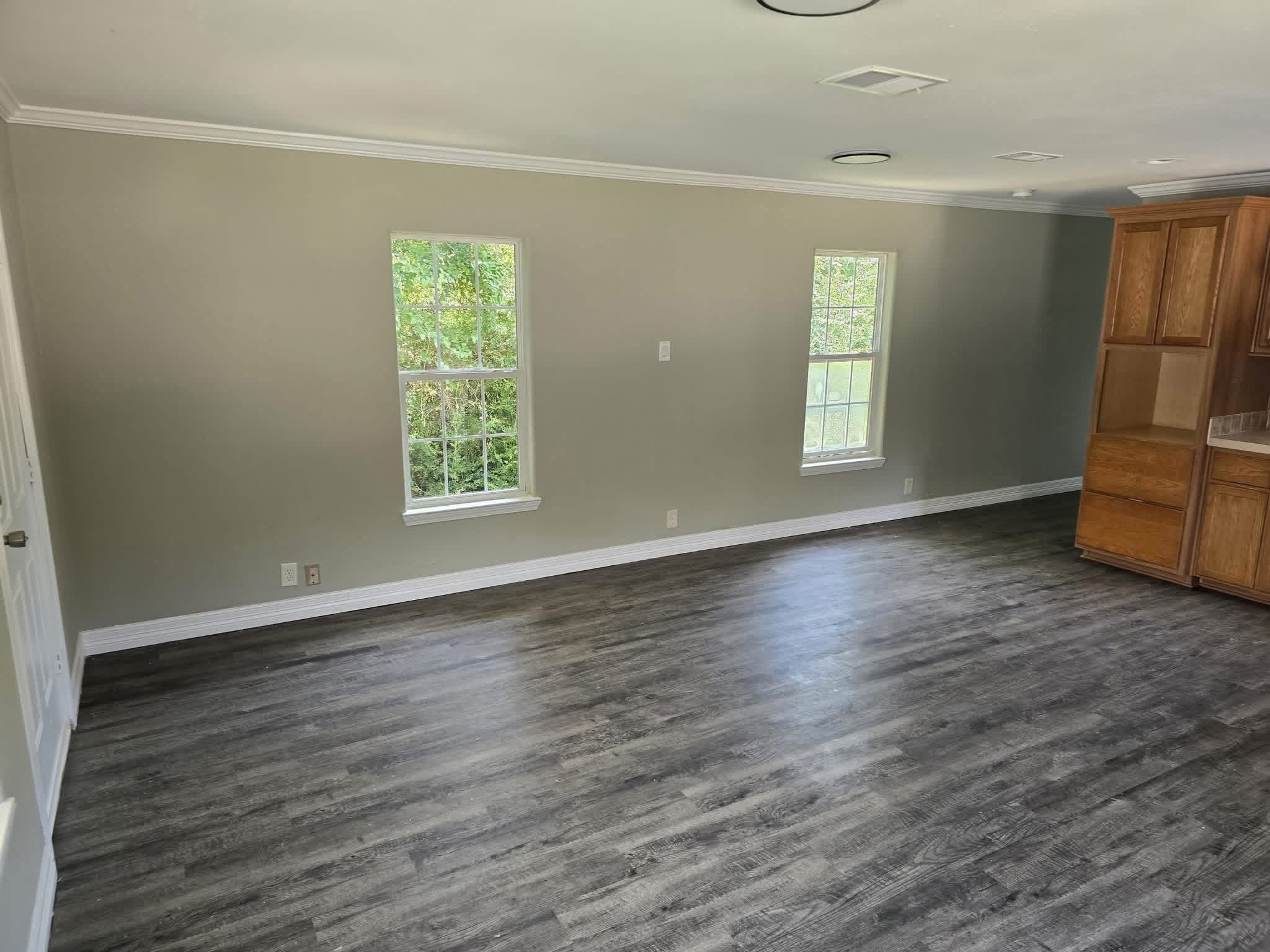 Spacious empty room with gray walls, dark wood flooring, two white-framed windows, and wooden cabinets creating a bright, modern, open atmosphere.