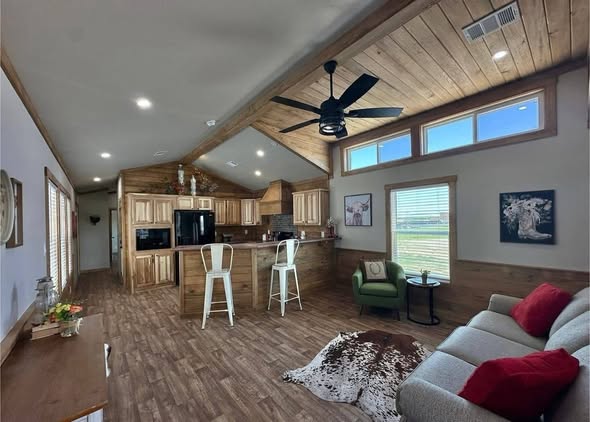 Modern luxury rustic-style park model home interior featuring a spacious open-concept living area with high vaulted wood-plank ceilings, a contemporary black ceiling fan, and energy-efficient clerestory windows. The sophisticated design showcases a gourmet kitchen with warm natural wood cabinetry, a breakfast bar with white industrial metal stools, and premium wide-plank flooring, while the cozy living room includes a neutral sofa with vibrant red accents, a cowhide rug, and Western-themed decor. This high-end interior captures a professional "full rank" SEO-optimized aesthetic, perfect for luxury prefab vacation cottages, custom-built small homes, or stylish accessory dwelling units (ADU) with a refined cabin feel.