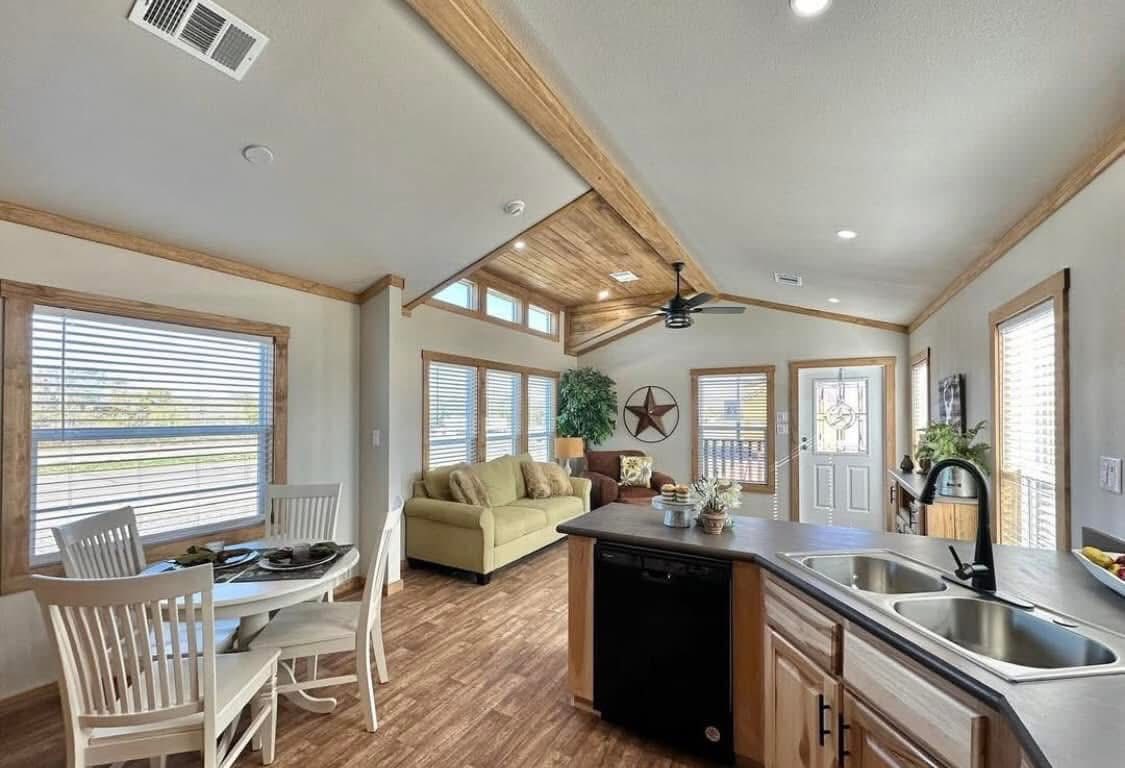 SEO-optimized alt text for a luxury park model tiny house featuring a navy blue board-and-batten exterior, a large gabled front porch with natural wood pillars and exposed timber beams, and a sleek black metal roof; the accompanying interior view showcases an open-concept living space with vaulted white shiplap ceilings, a modern kitchen with white cabinetry and stainless steel appliances, and a bright dining area with large windows, perfectly suited for high-ranking searches related to luxury mobile living, sophisticated vacation cottages, and innovative small-space architectural design.