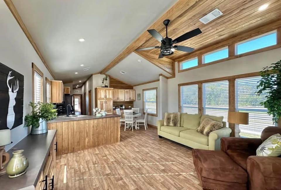 Spacious living room with wooden ceilings and floor, light beige sofa, brown armchair, and a ceiling fan. Bright kitchen and dining area in the background.