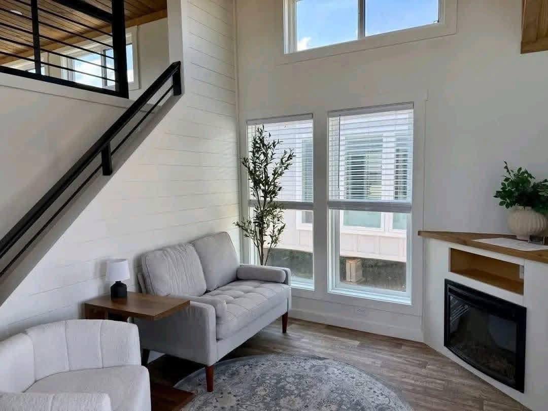 A cozy living room with a high ceiling features a gray sofa, white armchair, and a small table. Large windows provide natural light, and a fireplace adds warmth. A potted plant and rug enhance the airy, modern decor.