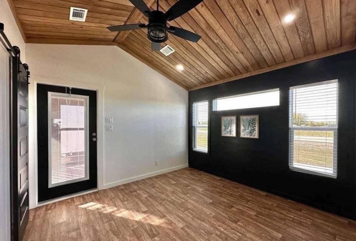  Gemini said Modern small home interior featuring a vaulted wood plank ceiling with a black ceiling fan, light oak vinyl flooring, and a bold black accent wall with white-trimmed windows, creating a stylish and space-efficient living area in a contemporary tiny house or modular home design.