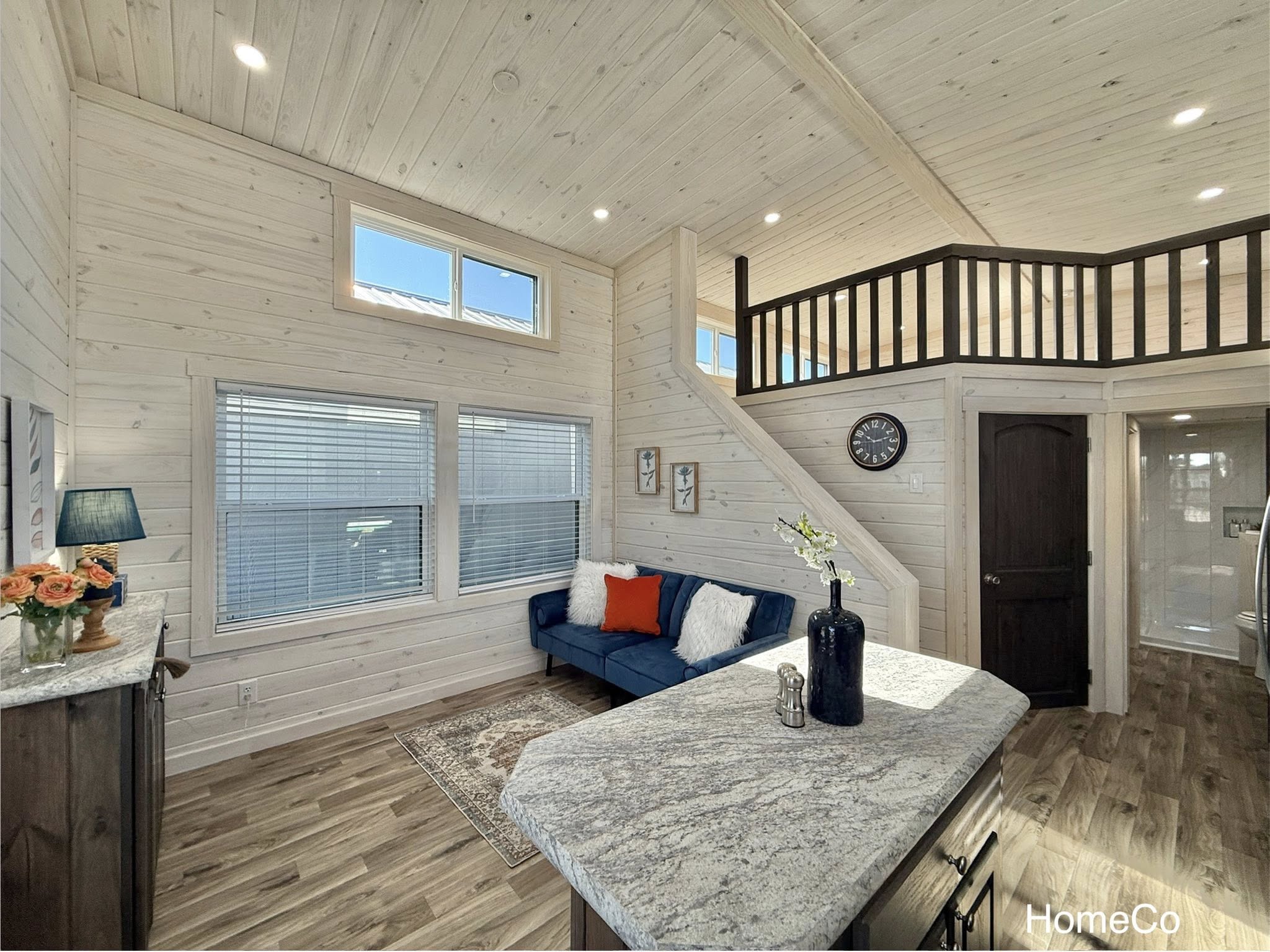 Bright, cozy living area with light wood paneling, high ceiling, and large windows. Features a blue sofa, stairs to a loft, and a rustic kitchen island.