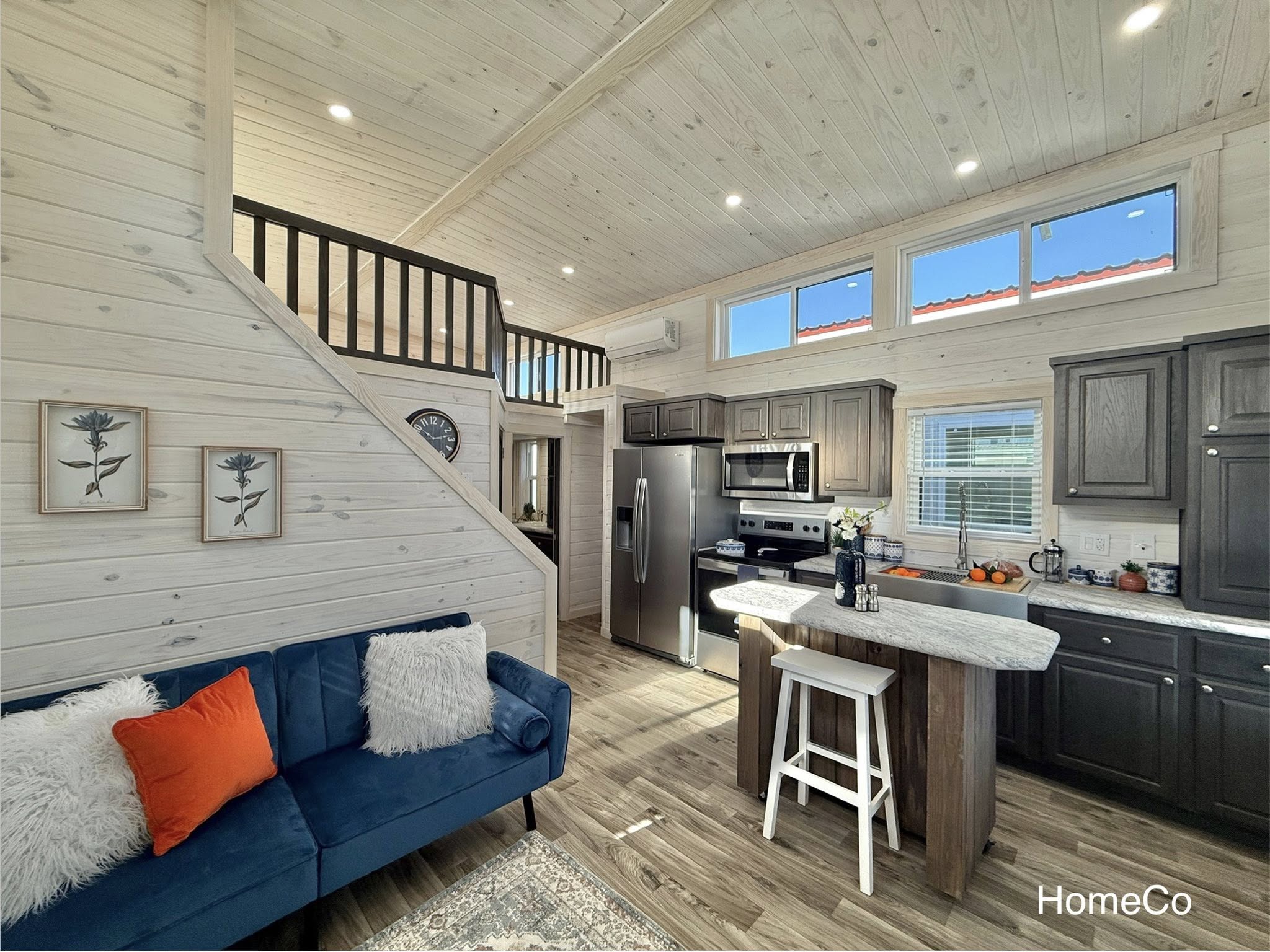A cozy tiny home interior with a loft above the kitchen, featuring wooden walls, a dark blue sofa with red and white pillows, and a modern kitchen with dark cabinets.