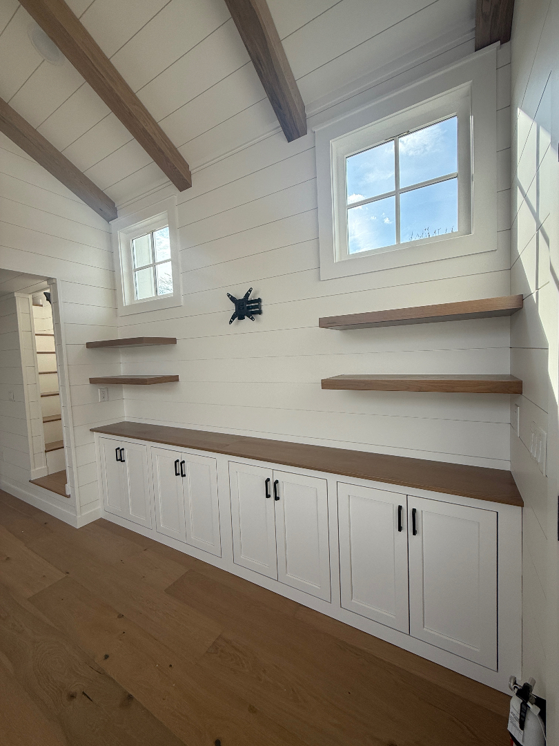 Modern farmhouse tiny house interior featuring white shiplap walls, vaulted ceilings with rustic wood beams, and built-in storage solutions including white shaker cabinets with natural wood countertops and floating wooden shelves. The space is illuminated by natural light from high square windows, showcasing a clean and minimalist aesthetic perfect for luxury park model cottages, mobile living, or small home organization and design inspiration.