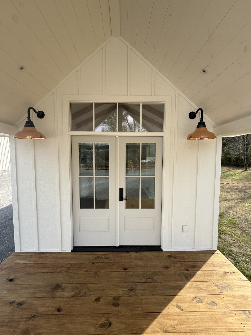 Luxury white modern farmhouse tiny house on wheels featuring a spacious covered front porch with a wood deck, copper gooseneck lighting, and double glass French doors. The professional park model cottage design includes a bright interior with a vaulted shiplap ceiling, open-concept kitchen with stainless steel appliances, a cozy living room with a fireplace, and a functional sleeping loft, perfect for high-end mobile living and upscale vacation rental marketing.