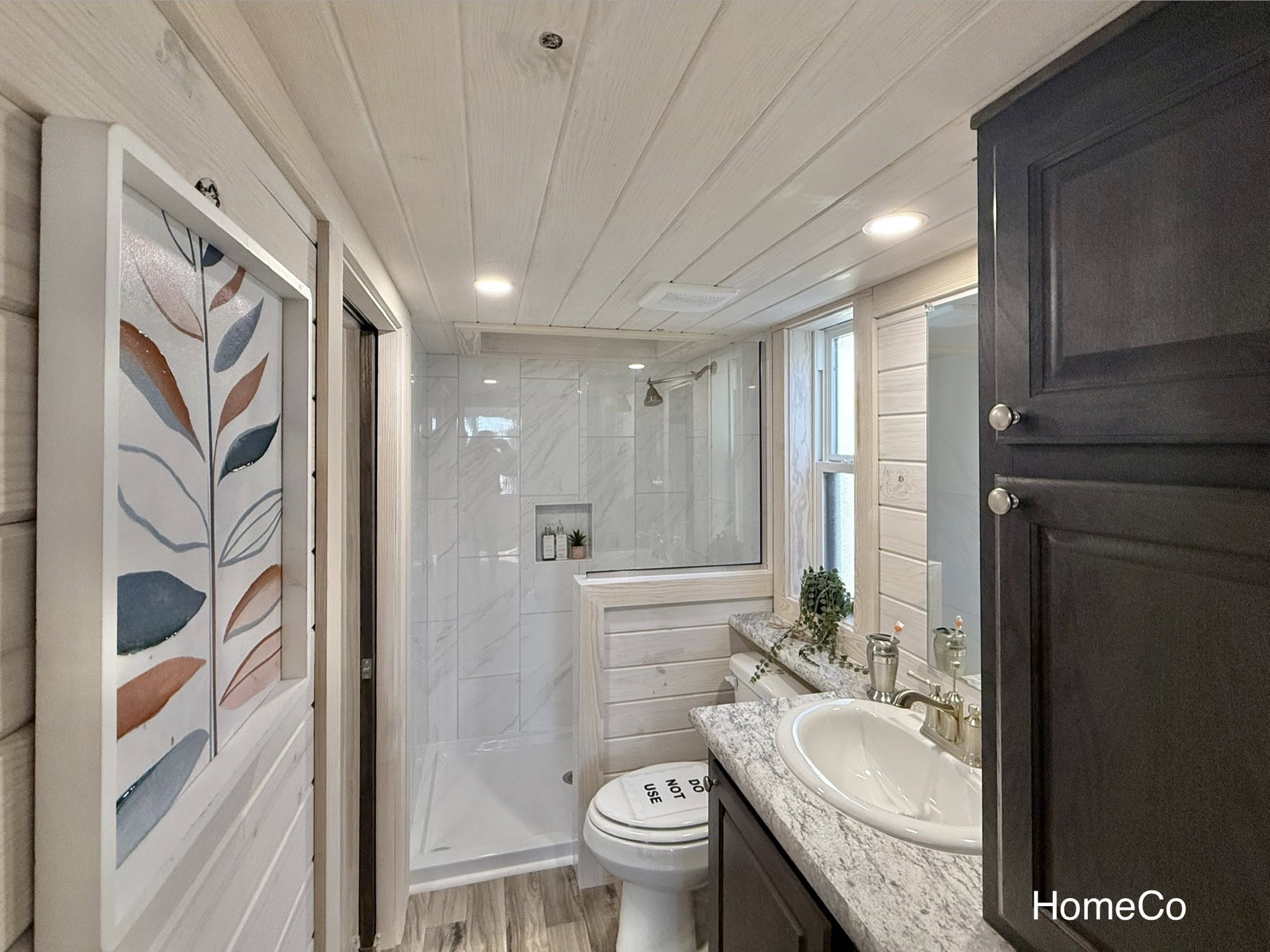 Modern bathroom with wood paneling, featuring a shower with glass door, sink with marble countertop, and decorative leaf artwork. Bright and cozy.