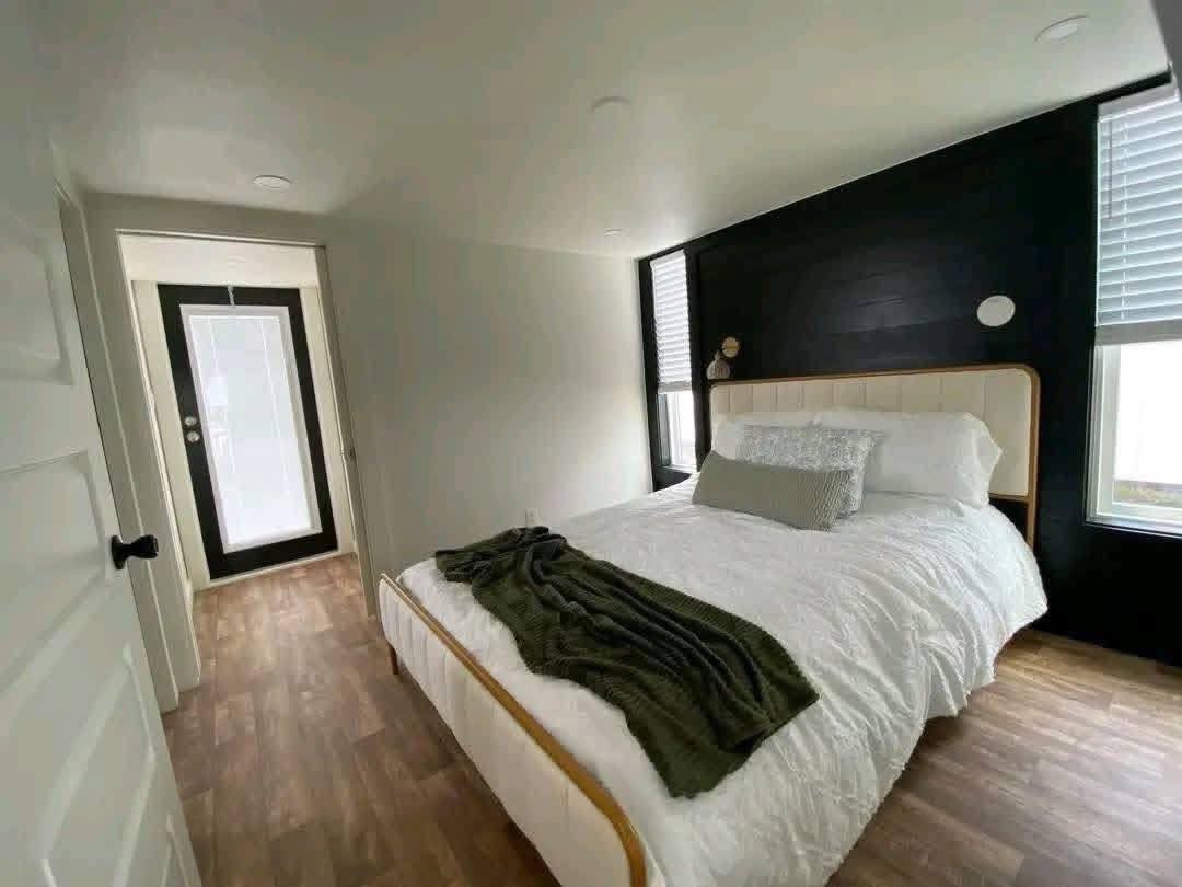 Minimalist bedroom with a black accent wall, white bedding, and wooden floor. A green throw is on the bed, with sunlight through two windows. Calm and cozy ambiance.
