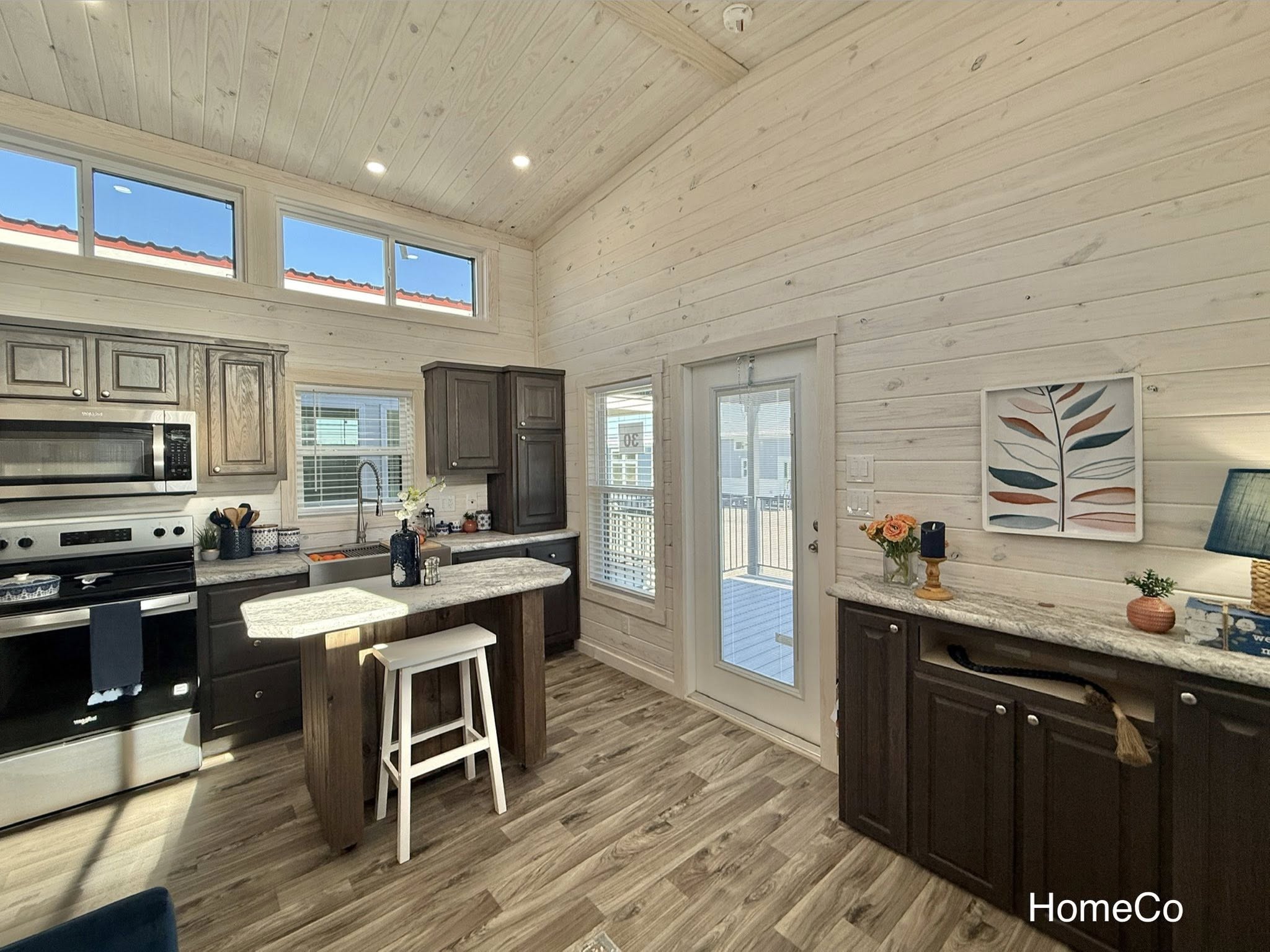 Bright kitchen interior with light wooden walls and ceiling, stainless steel appliances, and granite countertops. A small bar with stools and decorative elements like flowers and artwork enhance warmth.