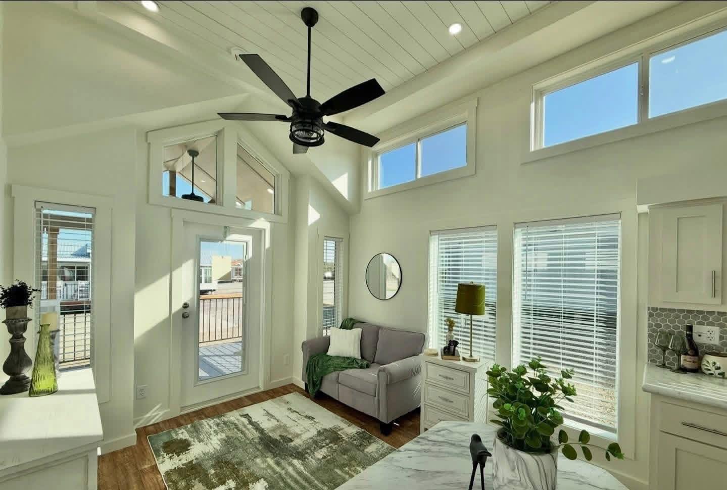Bright, cozy living room with high ceilings, a gray sofa, and green accents. Large windows, ceiling fan, and natural light create an airy feel.