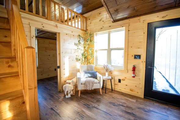 Cozy cabin interior with wood-paneled walls and floors. A plush white armchair by windows is adorned with a blanket and pillow. Natural light gives a warm ambiance.