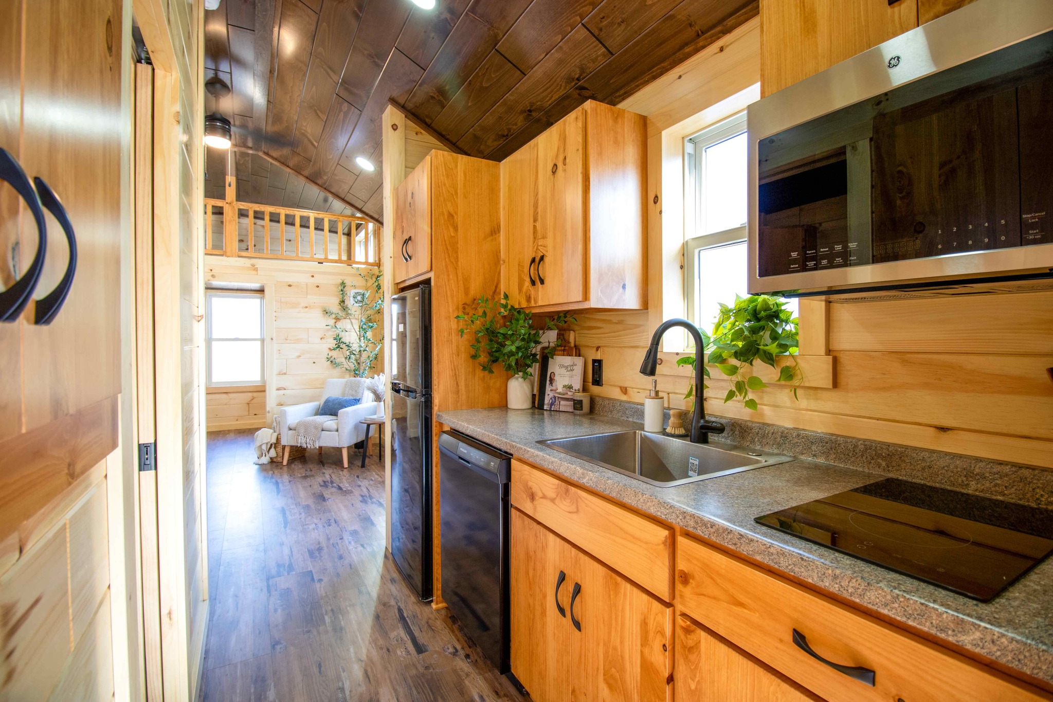 Warm, cozy kitchen in a wooden tiny house with pine cabinets, a dark countertop, green plants, and a small dining-living area in the background.
