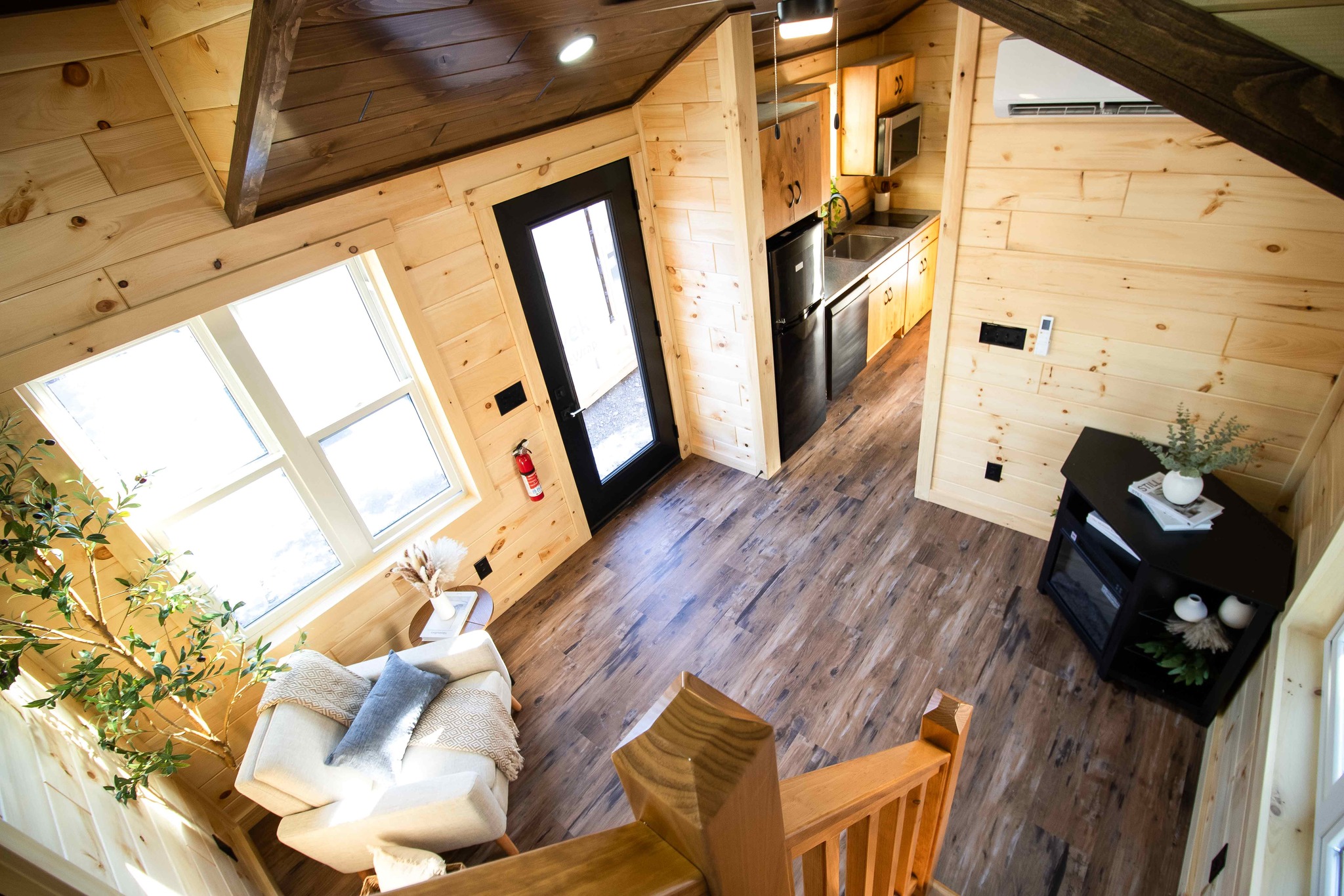 Cozy wood-paneled tiny home interior with a high ceiling. Features a doorway, a kitchen with stainless appliances, and a comfy chair near large windows.