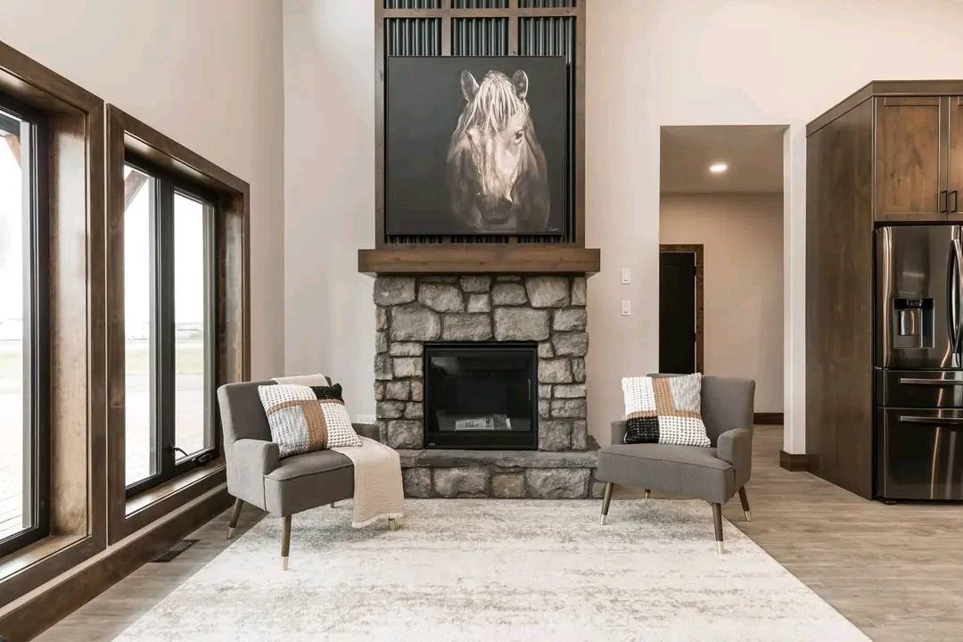 Stylish living room with a stone fireplace, horse portrait, and two grey chairs with pillows. Large windows offer natural light. Cozy and modern ambiance.