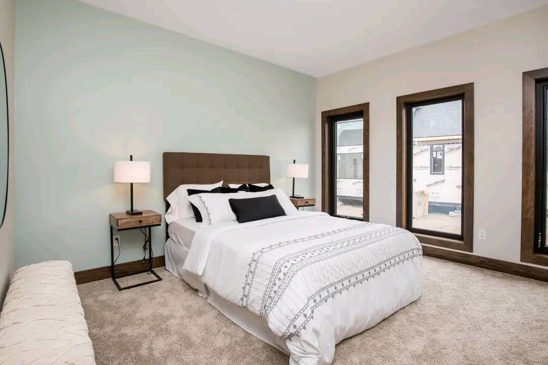 A serene bedroom with soft pastels. Features a bed with white and patterned linens, dual side tables with lamps, three large windows, and beige carpet.