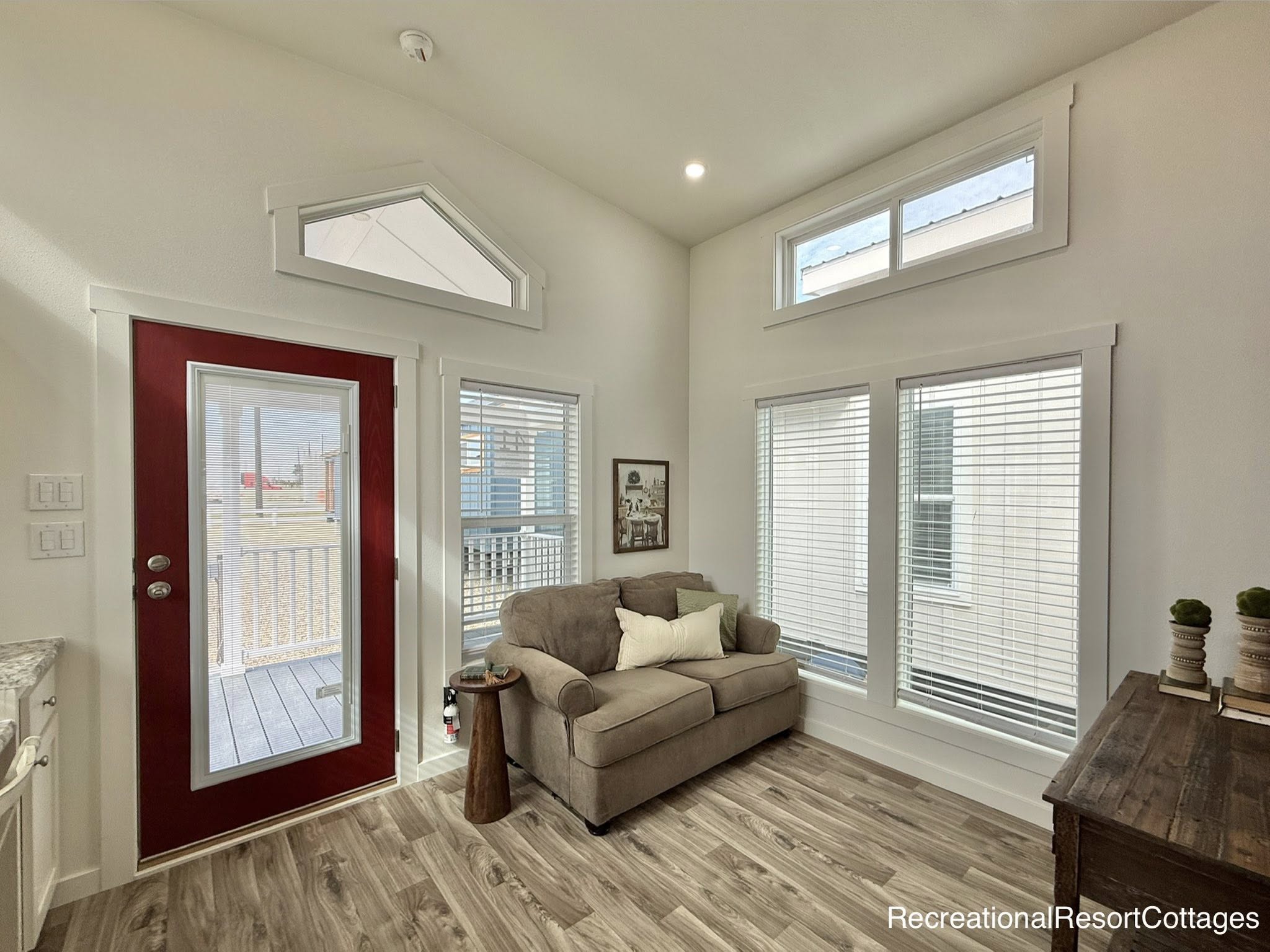 Cozy bright tiny home living room with high vaulted ceilings, white shiplap walls, large floor-to-ceiling windows with white blinds for abundant natural light, transom triangular window above red front door, neutral beige sofa with throw pillows, wooden side table, gray wood-look plank flooring, recessed lighting, modern minimalist farmhouse interior design in recreational resort cottage style – perfect for luxurious tiny house, vacation cabin, or small space living inspiration.