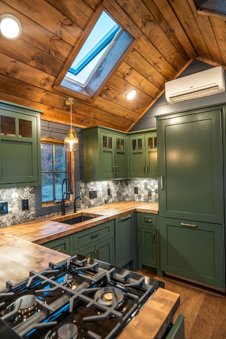 Modern luxury tiny home on wheels featuring dark charcoal vertical board and batten siding, a durable black metal gabled roof, and a stunning integrated front porch with natural cedar pillars and matching double wood-framed glass doors. The high-end architectural design is paired with a sophisticated interior showcasing a gourmet kitchen with deep forest green shaker-style cabinetry, rustic butcher block countertops, a professional-grade ZLINE gas range, and a dramatic vaulted ceiling with natural wood planks and integrated skylights. This premium "Denali" park model captures a refined rustic-modern aesthetic with hexagonal stone tile backsplashes, industrial-style lighting, and high-end wide-plank flooring, offering a perfect "full rank" SEO-optimized small-space living solution for luxury prefab houses or custom-built vacation retreats.