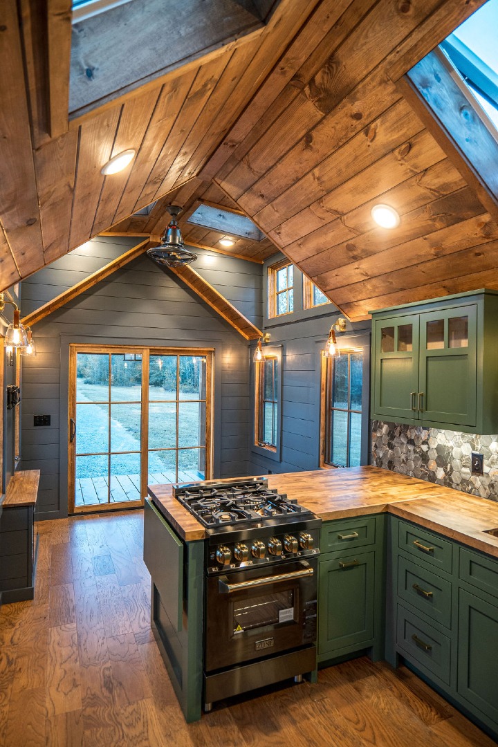 Modern luxury tiny home on wheels featuring dark charcoal vertical board and batten siding, a durable black metal gabled roof, and a stunning integrated front porch with natural cedar pillars and matching double French doors. The high-end architectural design is paired with an airy open-concept interior showcasing a gourmet kitchen with sage green shaker-style cabinetry, butcher block countertops, a professional-grade ZLINE gas range, and a sophisticated vaulted ceiling with natural wood planks and integrated skylights. This premium "Denali" park model captures a rustic-modern aesthetic with industrial-style lighting and high-end wide-plank flooring, offering a perfect "full rank" SEO-optimized small-space living solution for luxury prefab houses or custom-built vacation retreats.