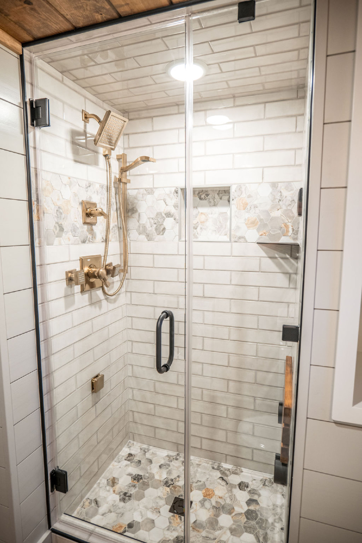 Modern luxury "Denali" tiny home walk-in shower featuring a frameless glass door with a matte black handle and high-end gold-finished fixtures, including a large rain showerhead and handheld sprayer. The sophisticated design showcases classic white subway tile walls accented by a decorative marble hexagonal tile inlay and a matching hexagonal stone tile floor, creating a bright, spa-like aesthetic perfect for full-rank SEO-optimized high-end prefab living, custom tiny house bathrooms, or luxury vacation cabin interiors.