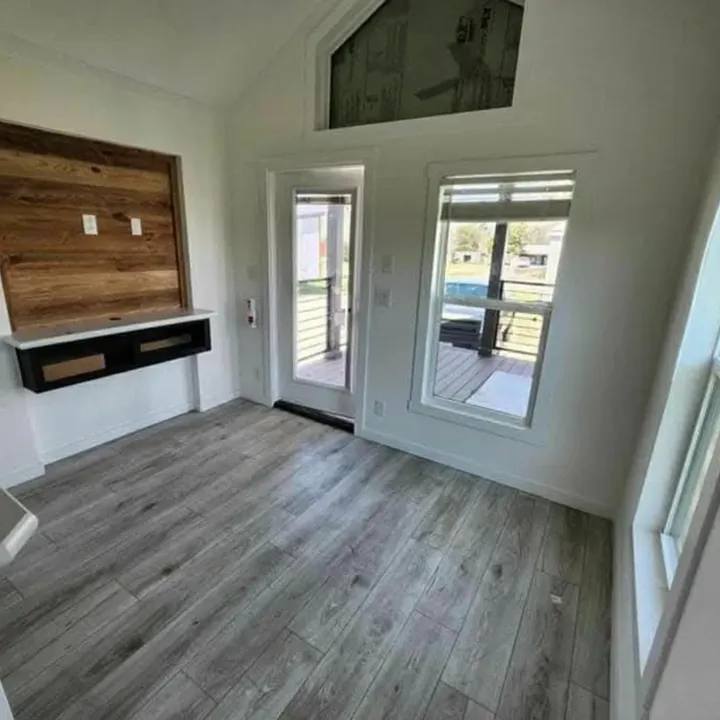 Modern luxury tiny home interior featuring a bright open-concept living area with premium grey wood-look flooring and crisp white walls, showcasing a rustic wood-plank accent wall with a built-in floating media console and a large glass entry door with a matching transom window that floods the minimalist space with natural light for a sophisticated move-in ready turn-key residential design.